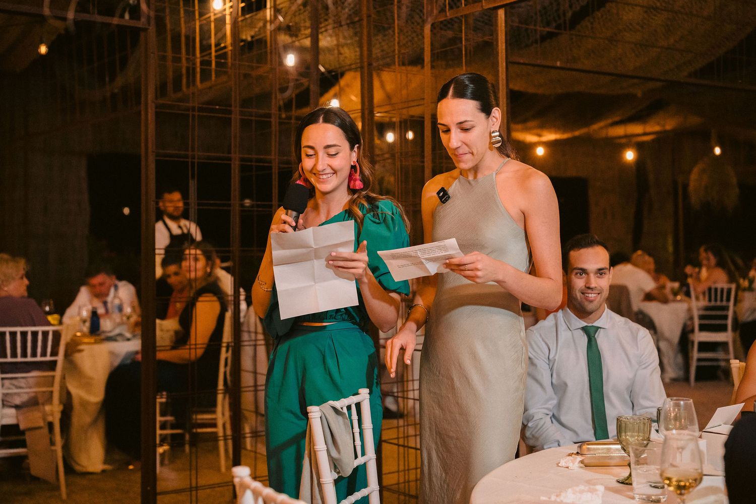 Two guests deliver speeches from papers while standing at an elegant indoor wedding reception with warm lighting.