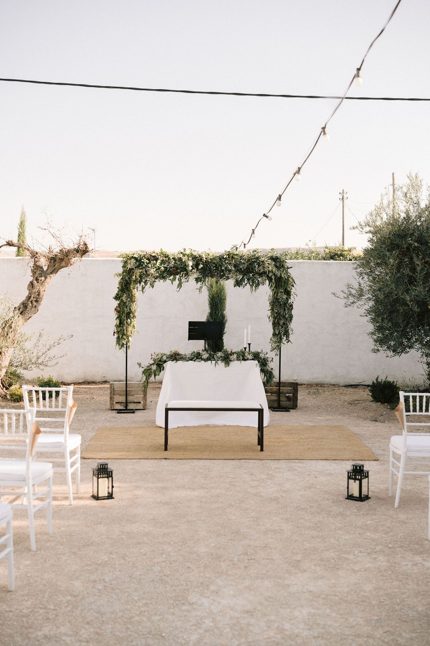 Minimalist outdoor wedding ceremony setup with greenery arch, white chairs, and lanterns against a white wall.