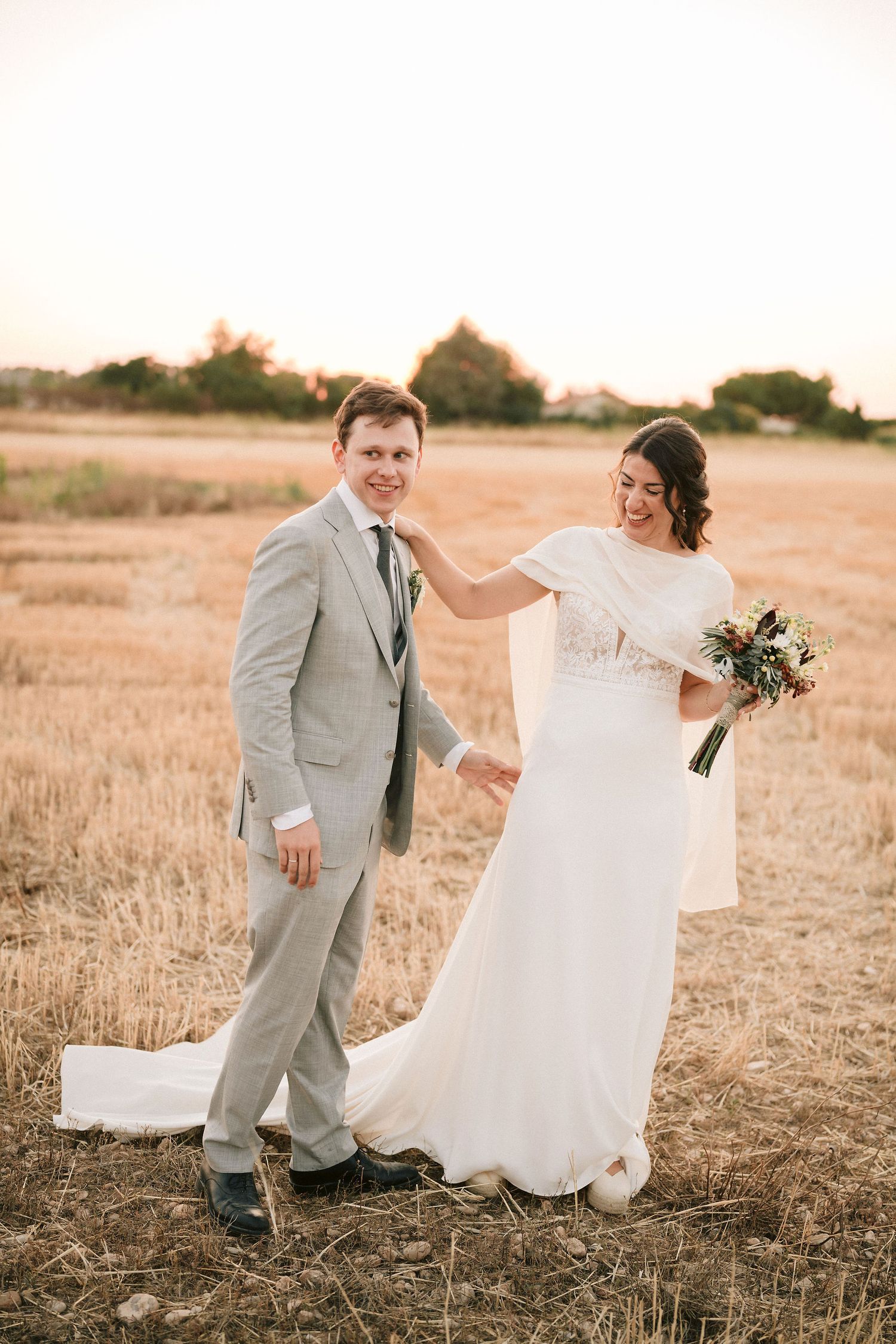 A bride in a flowing white dress and groom in a light gray suit share a candid moment in a golden field at sunset.