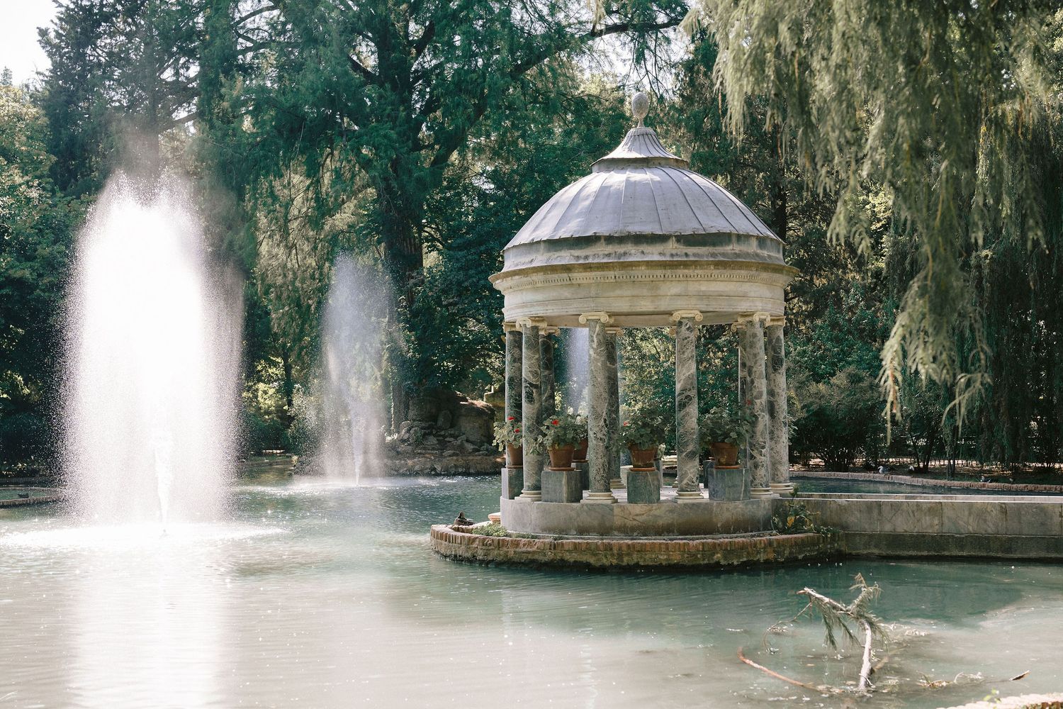 A serene garden fountain sprays water amid lush green foliage and hanging willow trees on a misty morning.