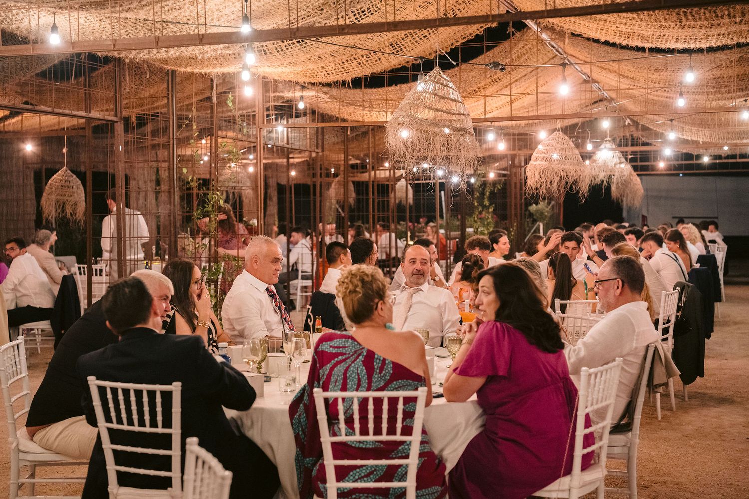 Wedding reception guests seated at tables beneath warm string lights in a rustic indoor venue during evening celebration.