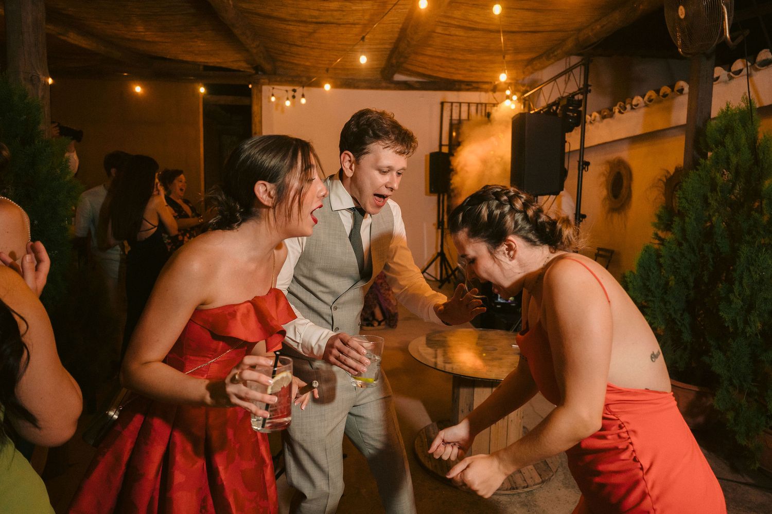 Wedding guests in red dresses dance with a person in a gray suit during a festive reception under warm lighting.