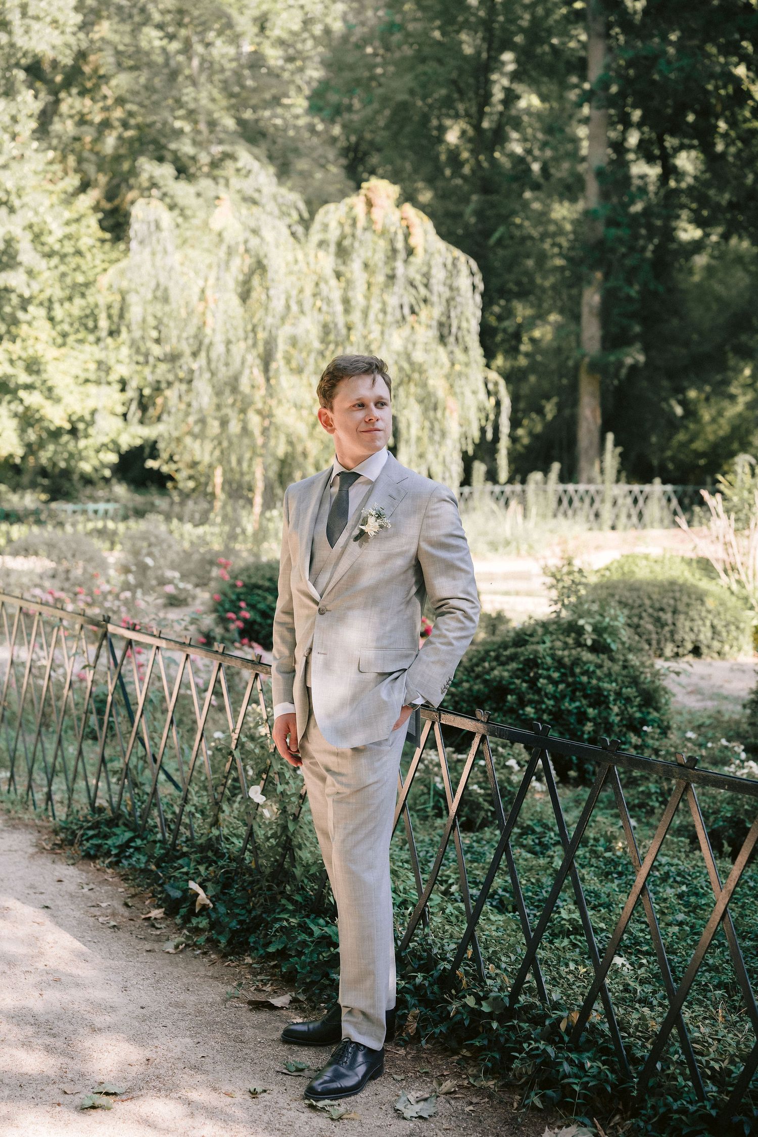 A person in a light gray suit poses outdoors along a wooden fence surrounded by lush greenery and garden foliage.