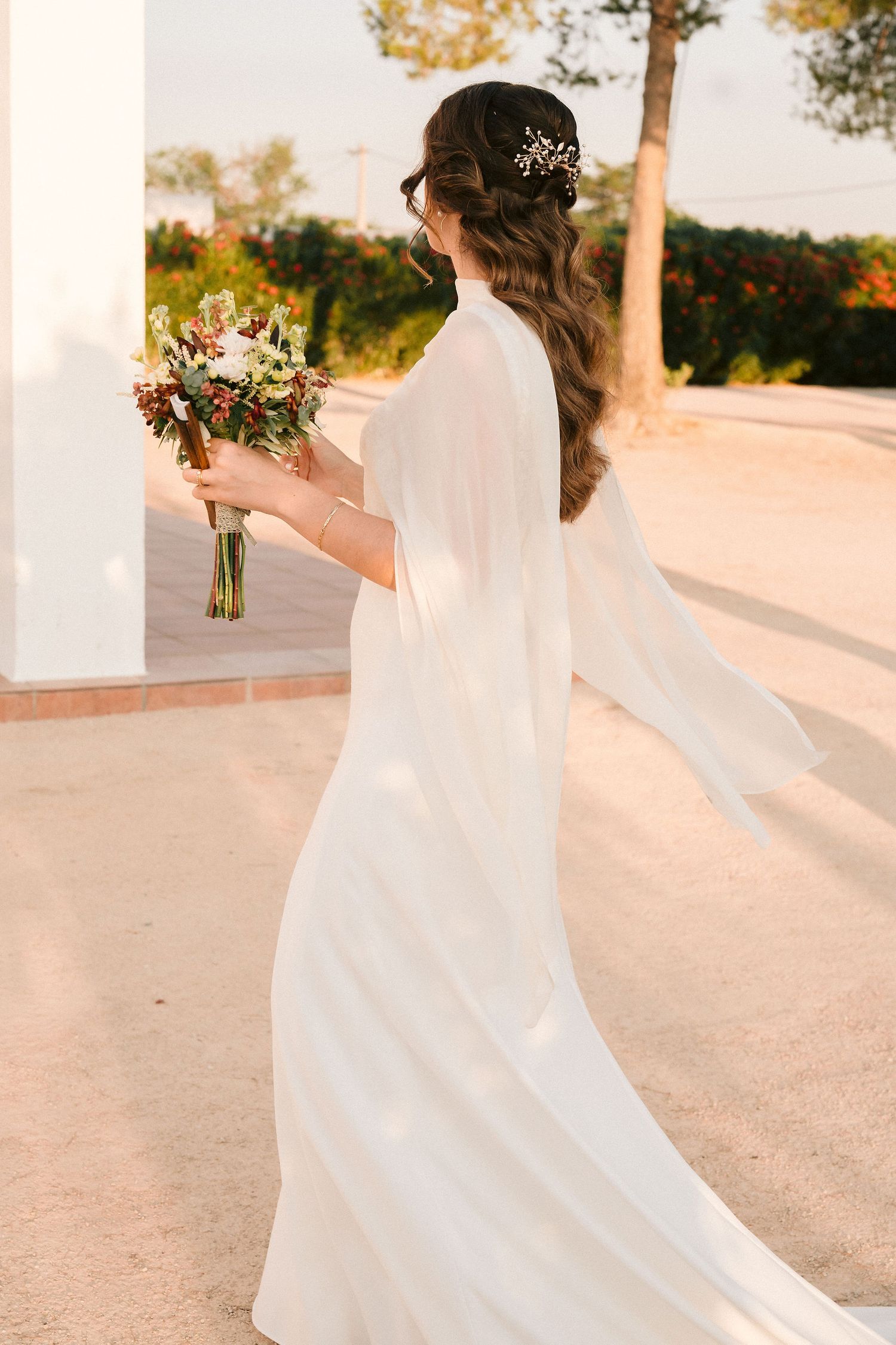 A bride in a flowing white dress with long sleeves holds a colorful bouquet while walking outside during golden hour.