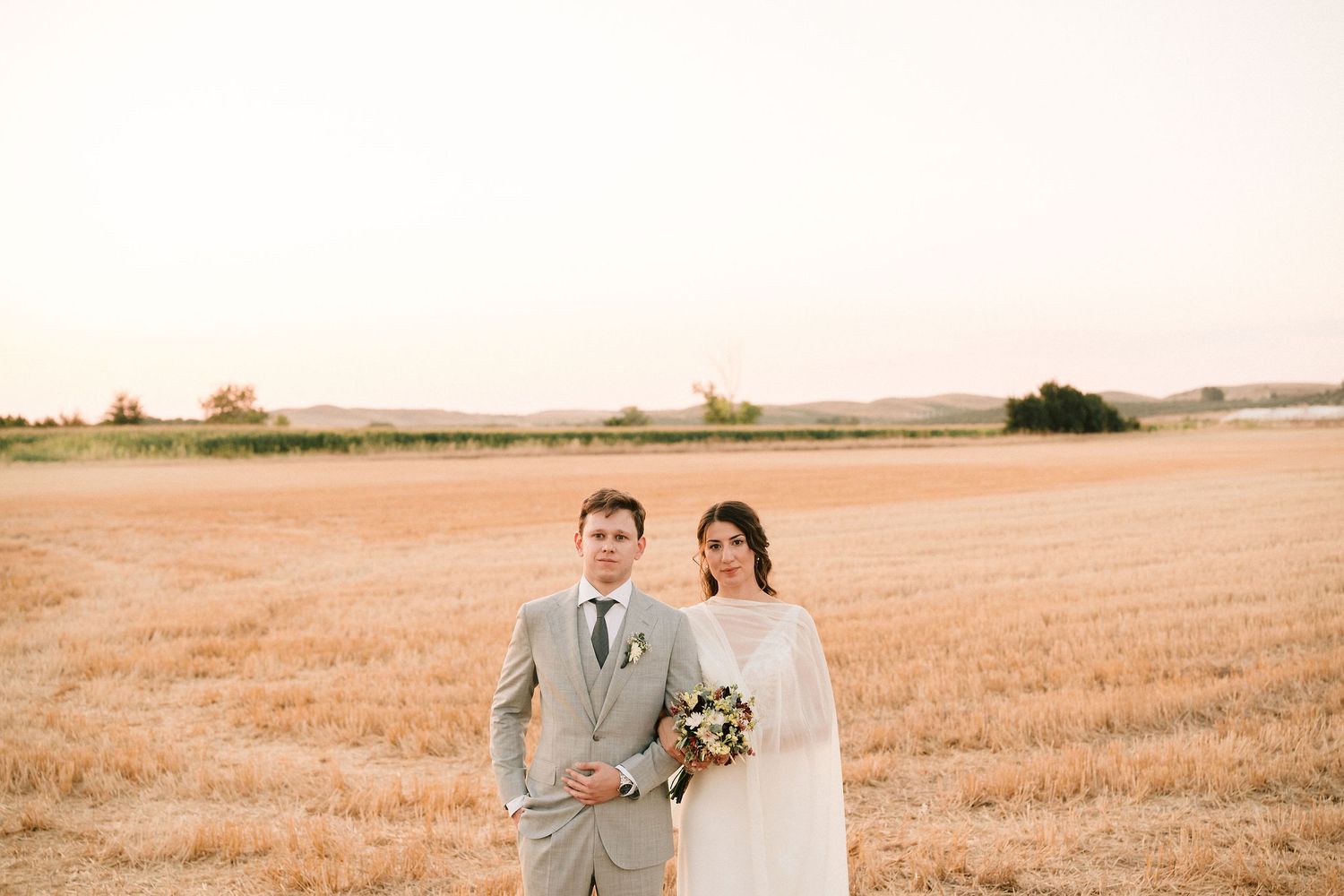 Newlywed couple walks hand-in-hand through a golden wheat field at sunset.