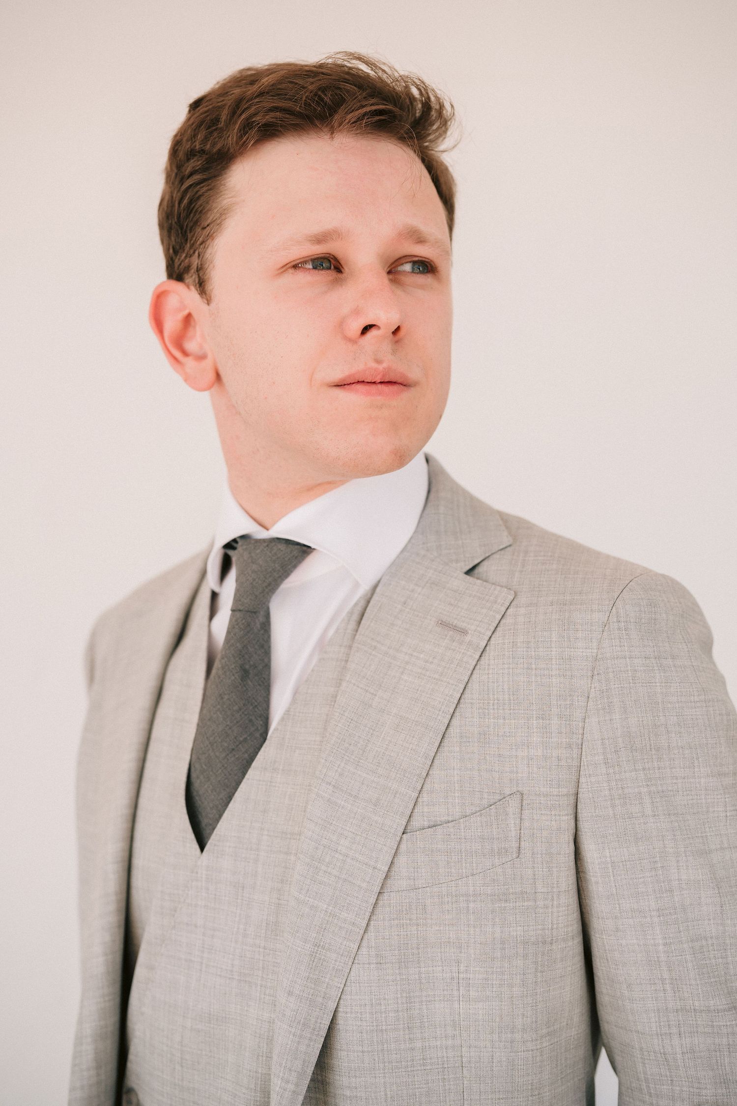 A person wearing a light gray three-piece suit with a dark tie poses against a neutral background.