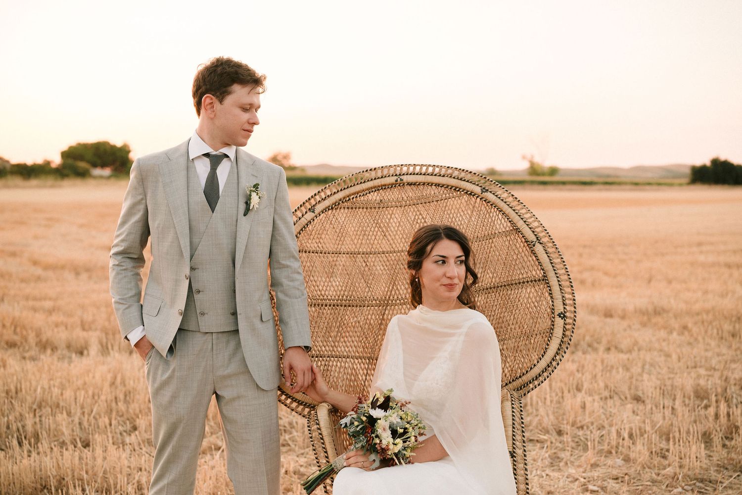 Romantic wedding couple poses together in wheat field with rattan peacock chair against golden sunset.