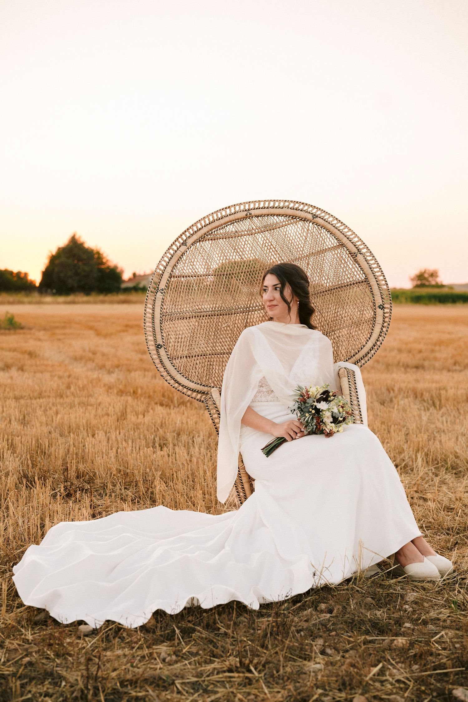 Wedding photography of a bride lounging on a wicker chair in a golden wheat field at sunset.