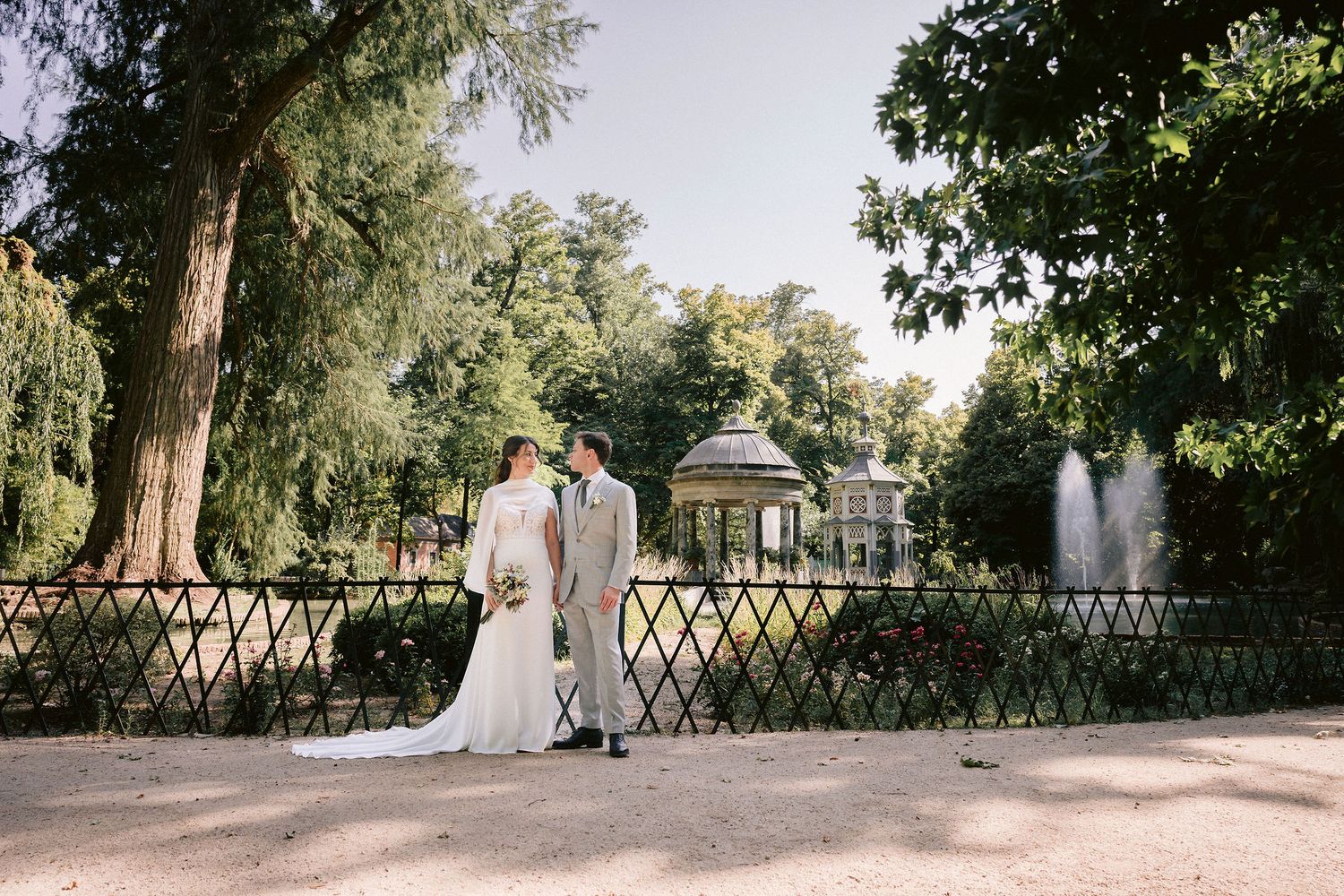Wedding couple walks hand in hand through lush garden pathway with beautiful natural lighting and greenery.