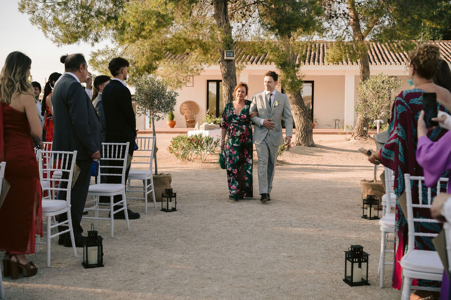 Wedding ceremony processional in an outdoor garden venue with lantern-lined aisle and white chairs for guests.