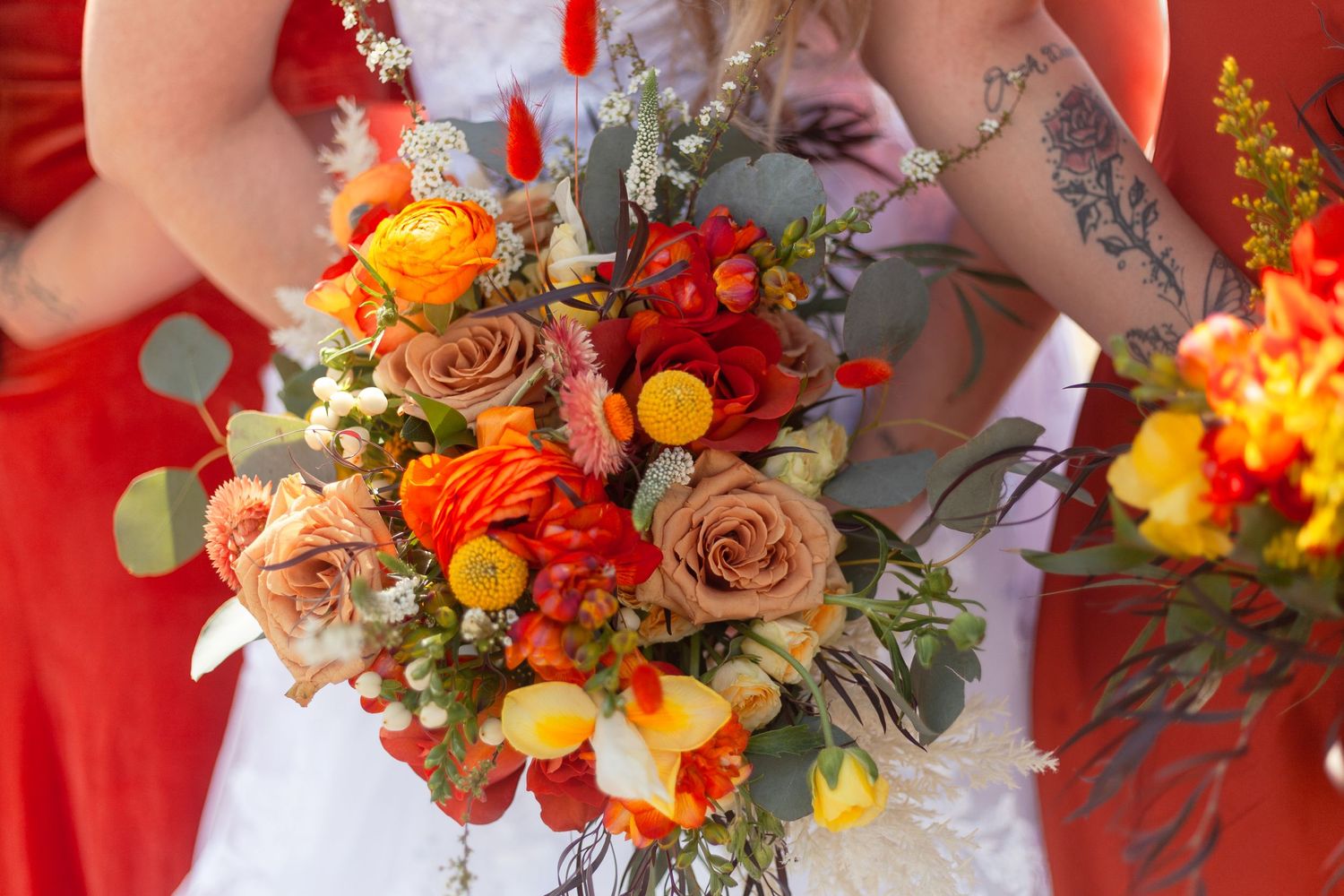 Vibrant fall-colored wedding bouquet with orange and red flowers held by bridesmaids.