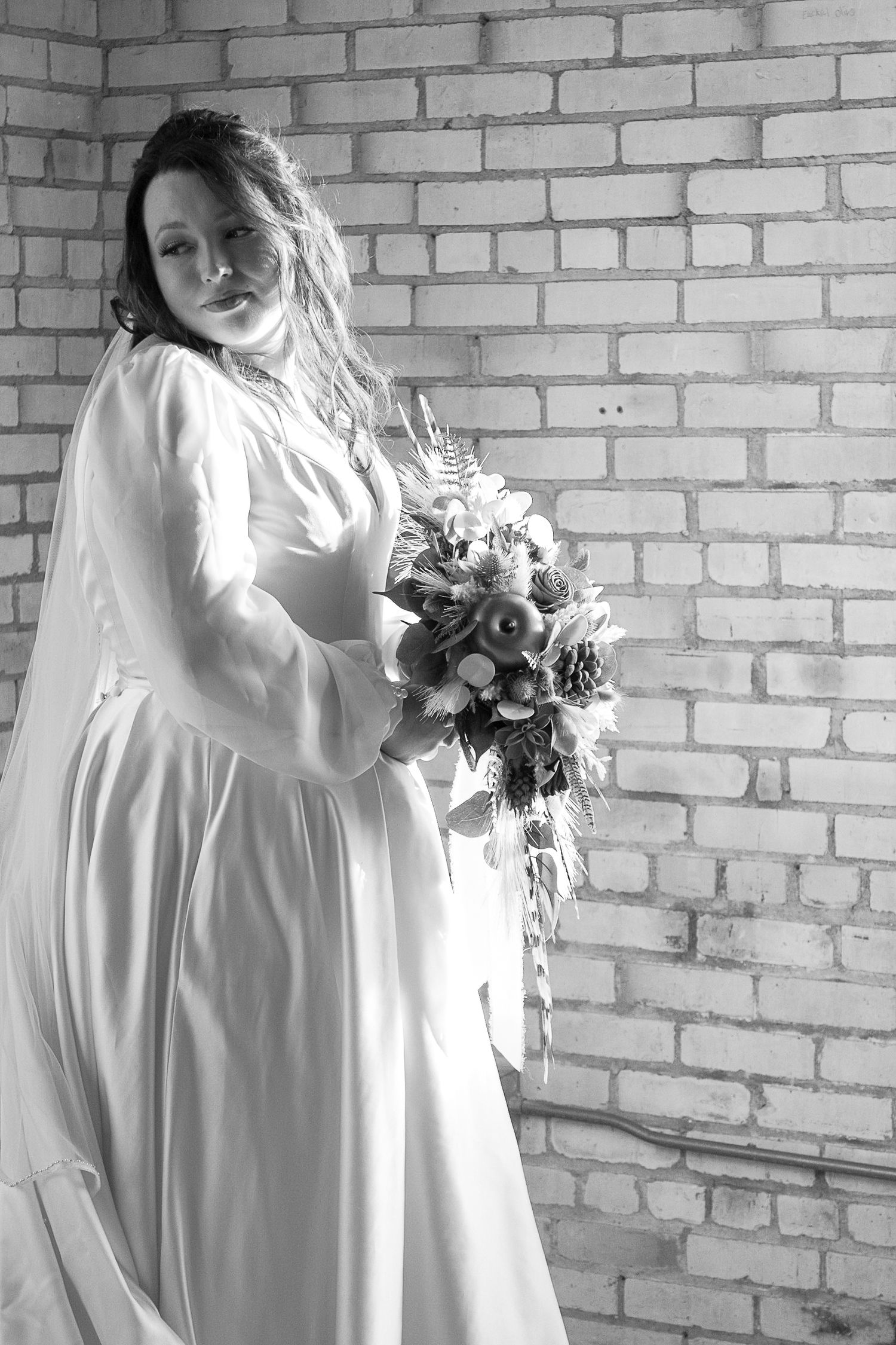 An artistic black and white bridal portrait against a brick wall features a flowing wedding dress.