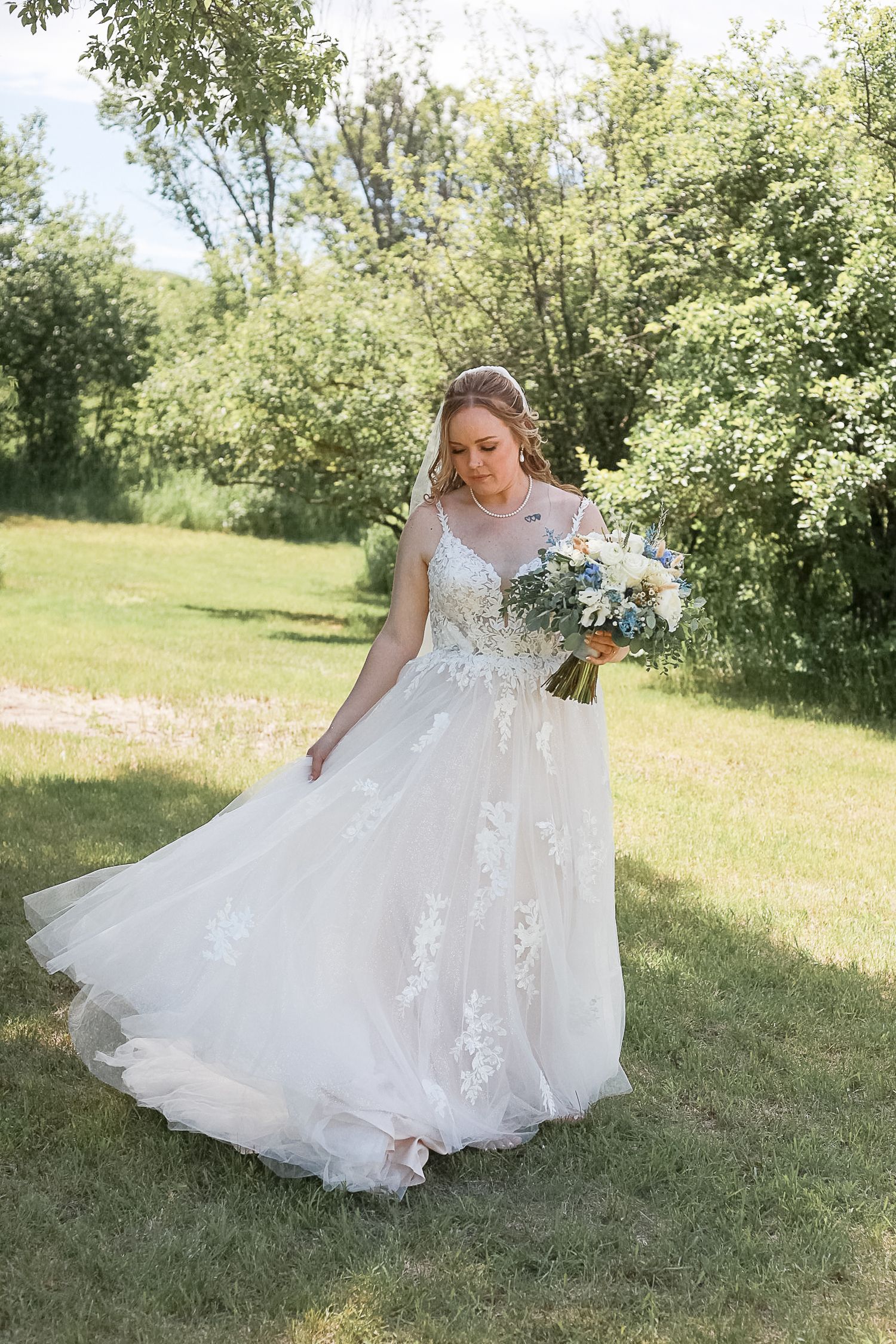 A bride in a delicate lace wedding gown holds a bouquet while standing in a sunlit garden setting.