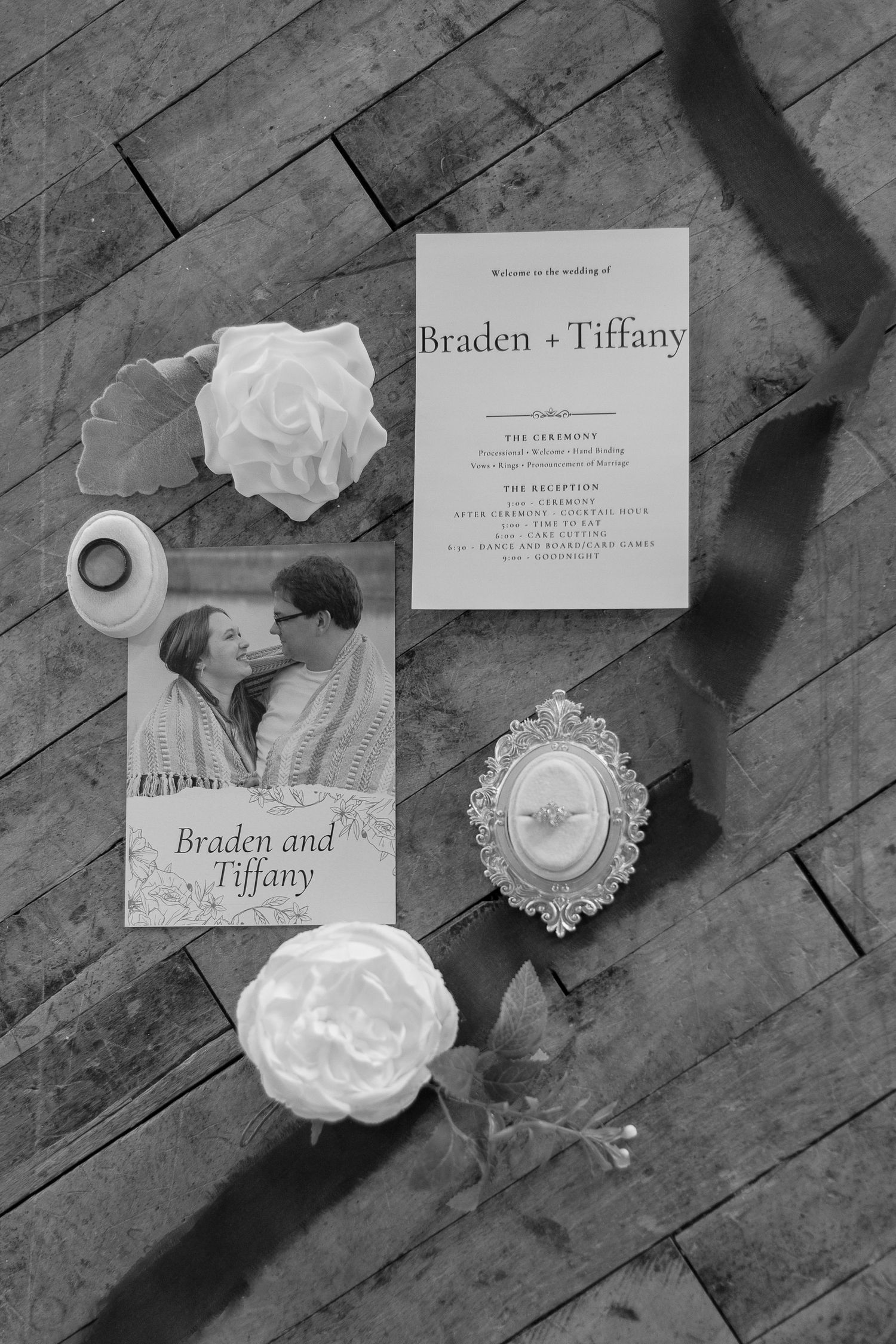 Black and white wedding invitation with decorative elements and floral accents on wooden floor.