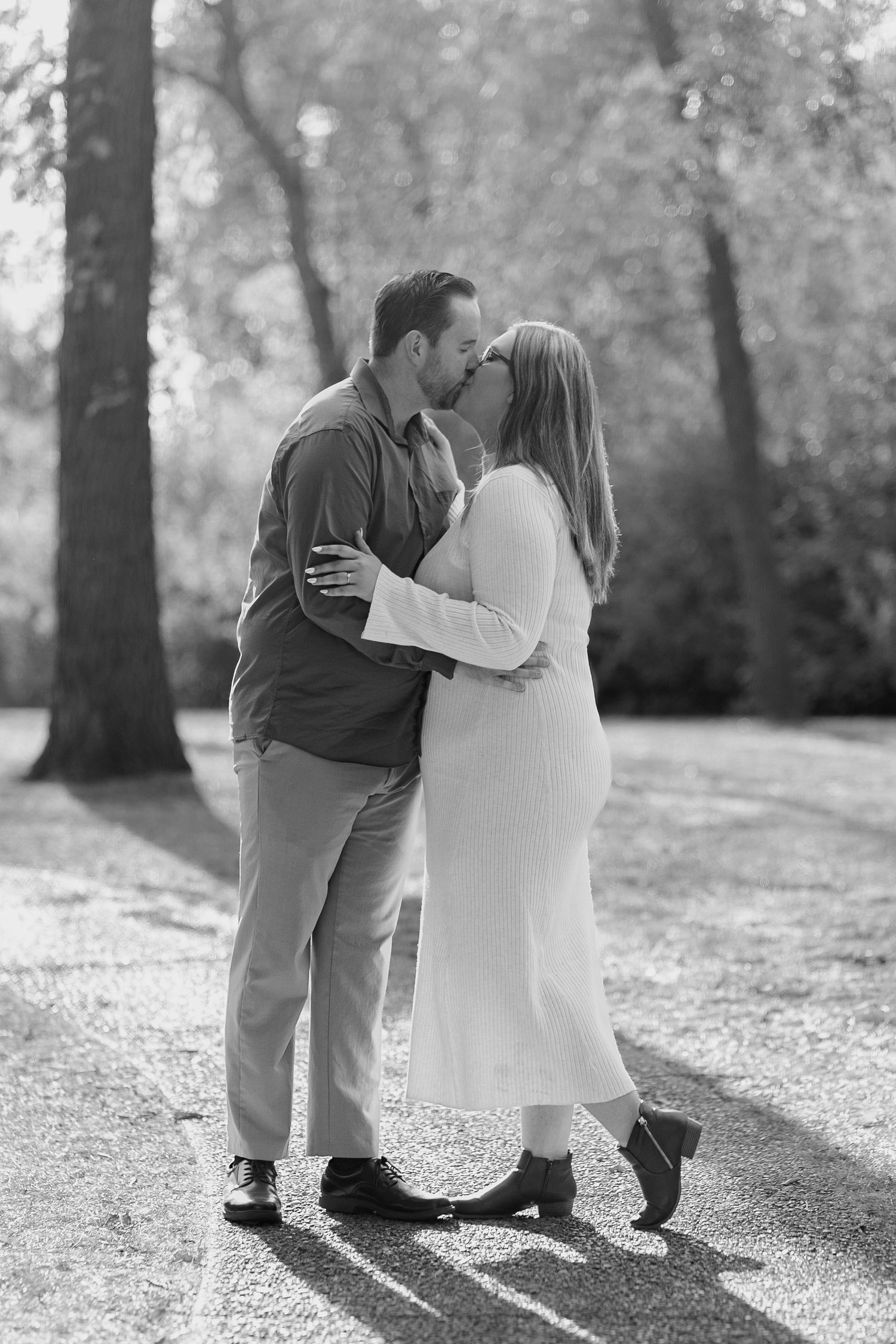 A black and white engagement photo shows a tender moment between a couple on a tree-lined path.