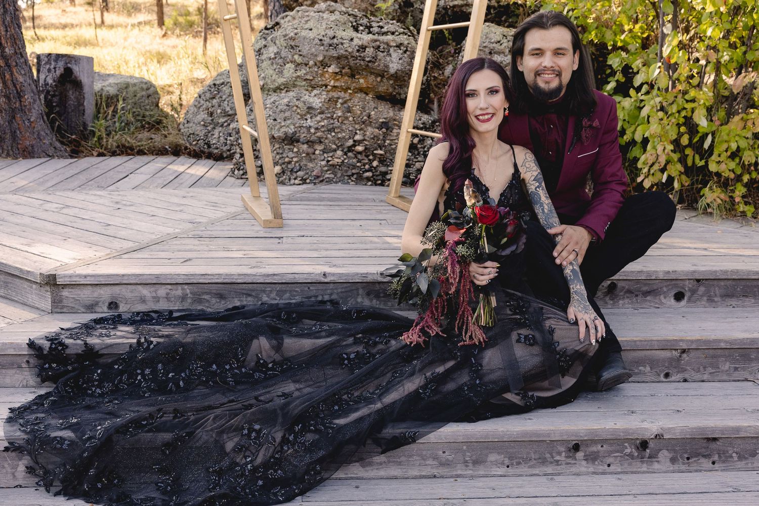 Couple in gothic wedding attire posing on wooden deck with dramatic black dress.