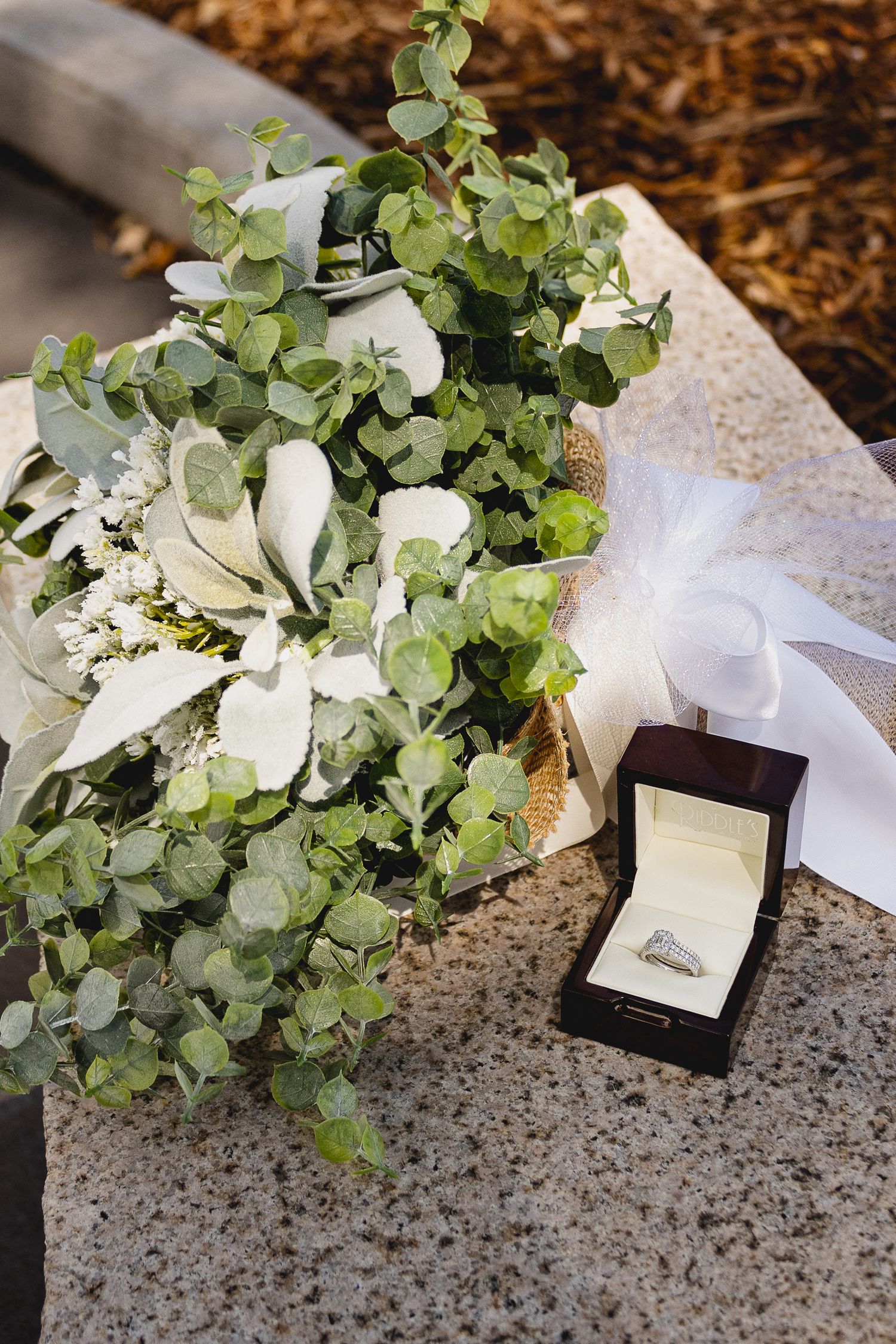 Wedding bouquet with eucalyptus leaves and white flowers next to an engagement ring box on stone surface.