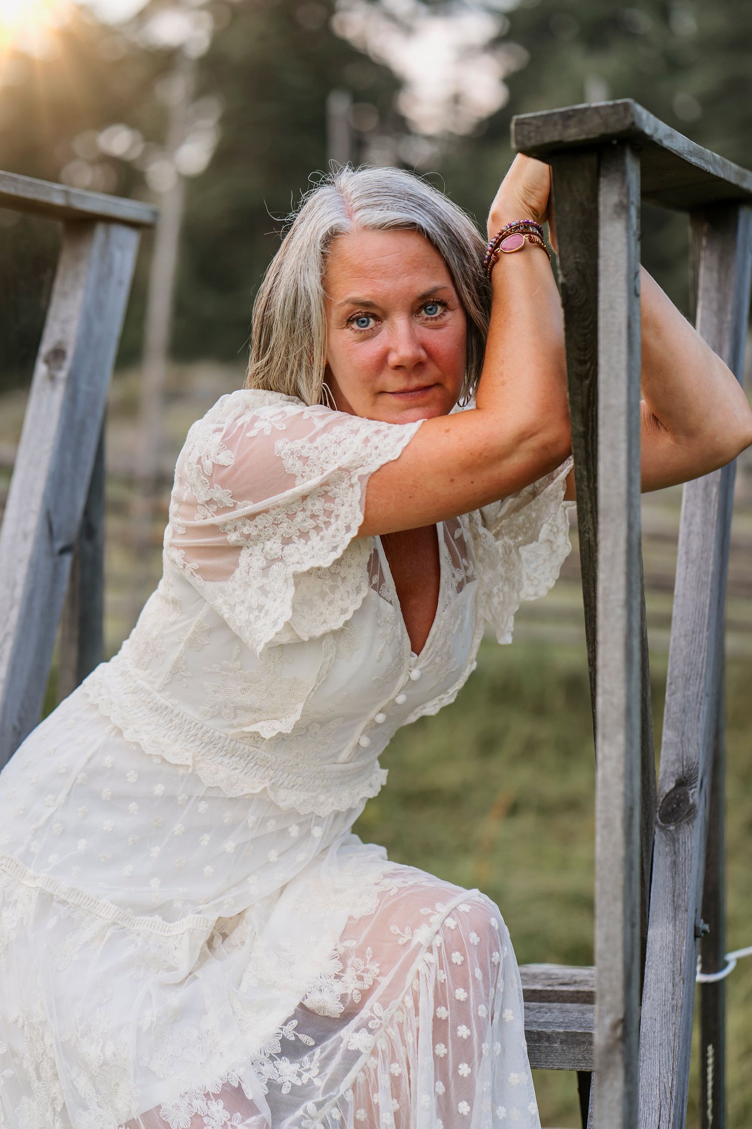 Woman in white lace dress poses playfully on rustic wooden stairs outdoors.
