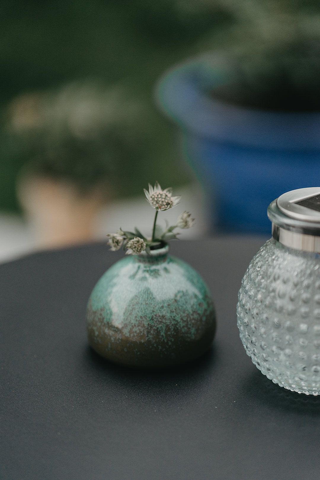 A delicate white wildflower in a small turquoise ceramic vase sits beside a textured glass jar on a dark surface.