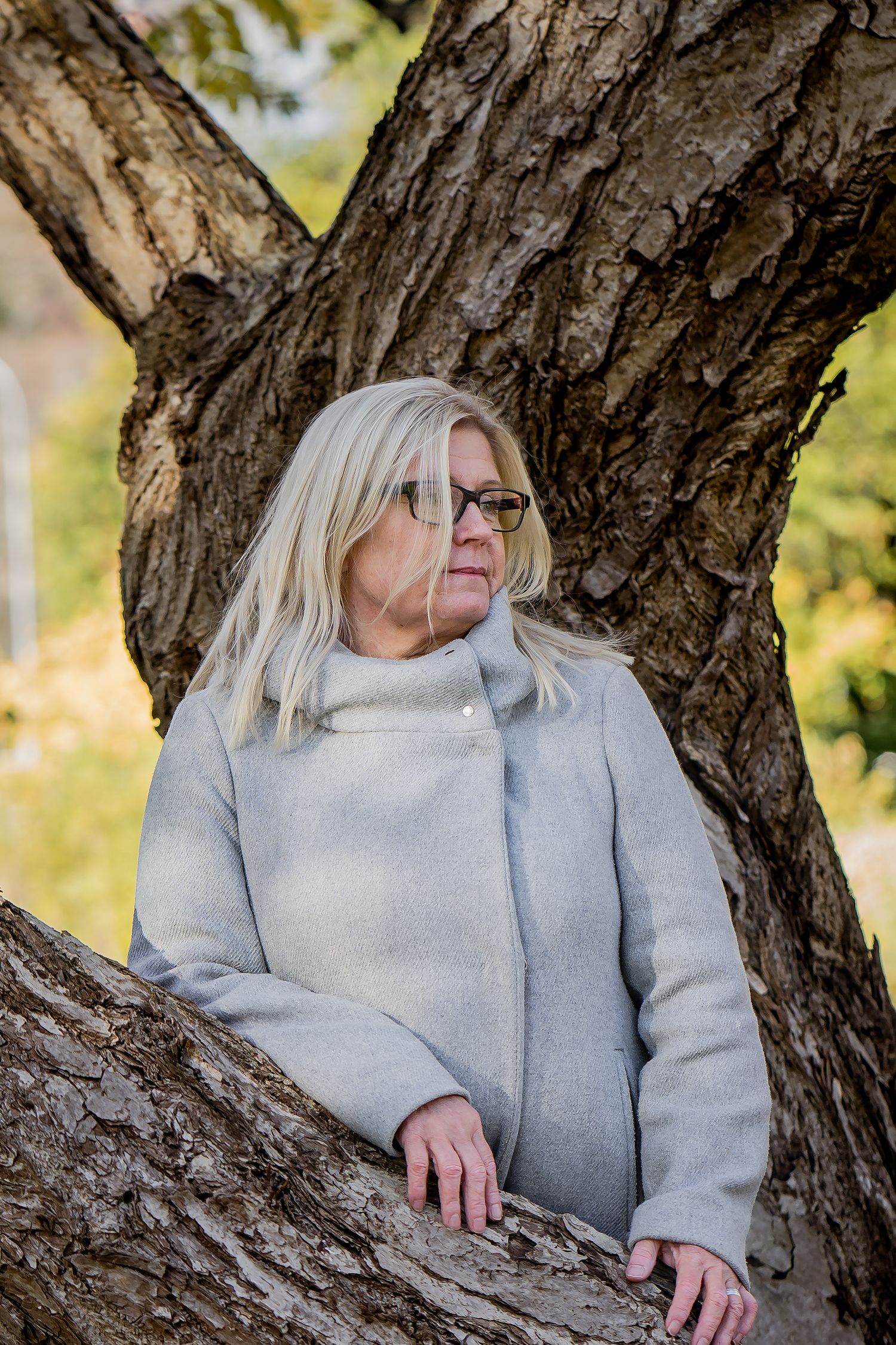 Woman in gray winter coat and glasses sitting in a rustic tree trunk on a sunny autumn day.