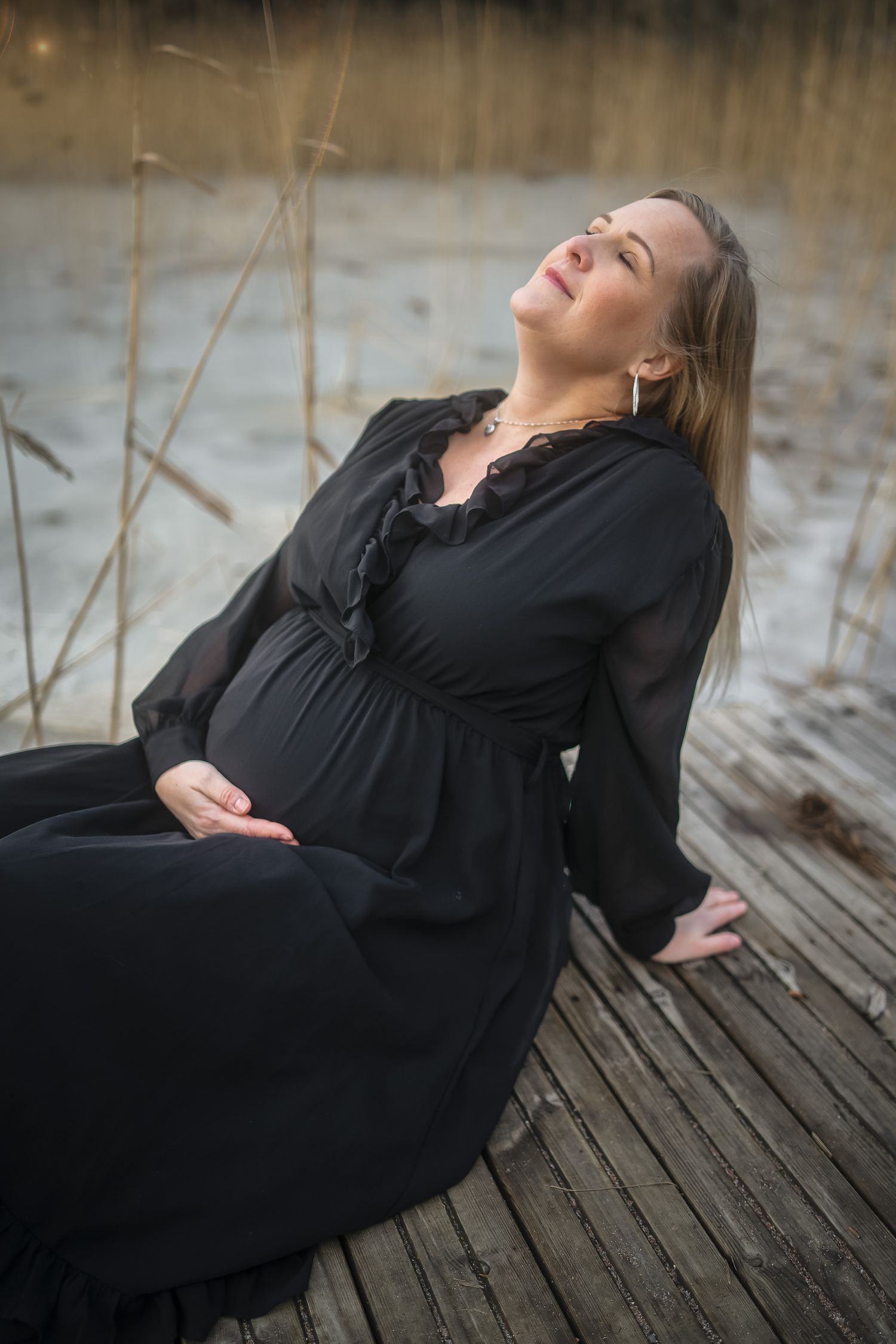Expectant mother in black dress sits peacefully on wooden dock by waterfront at sunset.