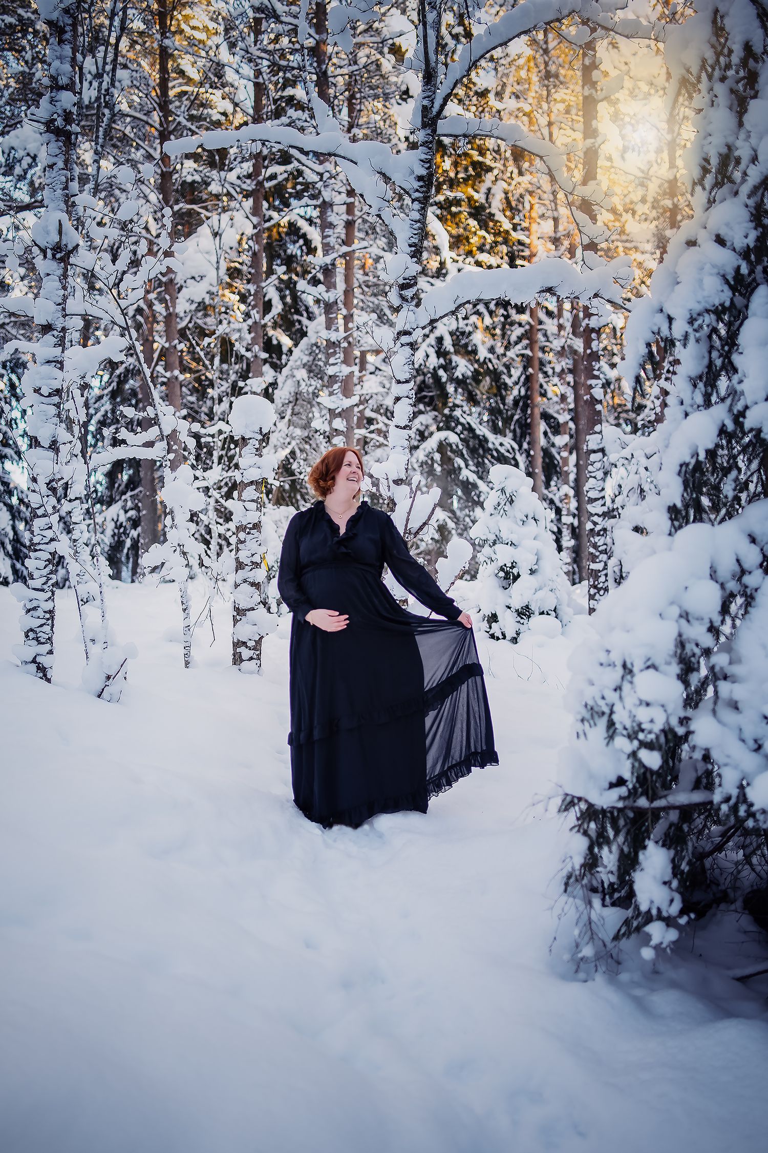 Person in black dress standing in snowy winter forest with sunlight filtering through trees.