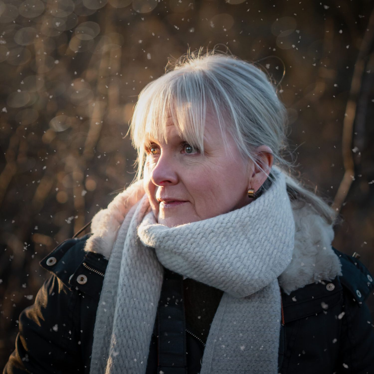 Person in knit scarf and winter coat during snowfall with bokeh effect in background.