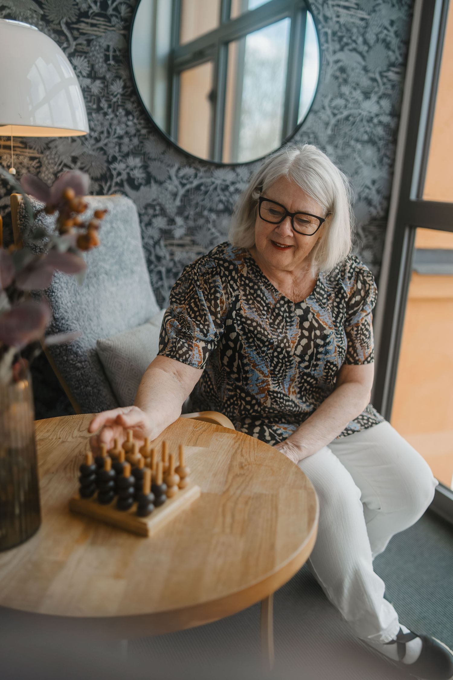 Person playing chess at round wooden table with floral wallpaper and modern decor.