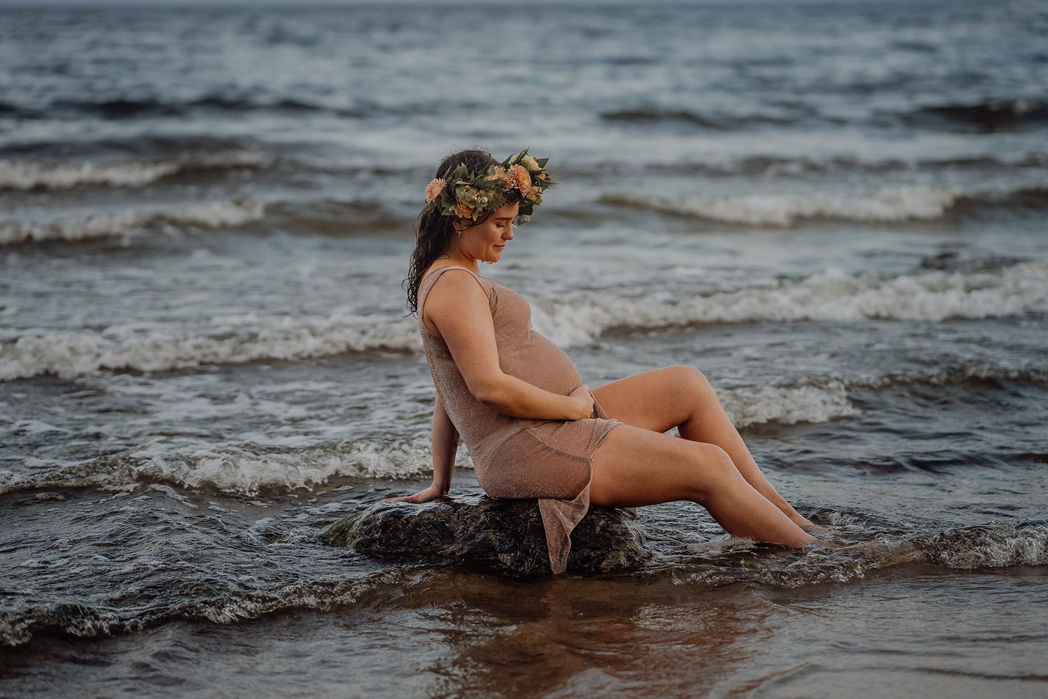 Person wearing floral crown sits on rock in ocean waves during sunset photoshoot.