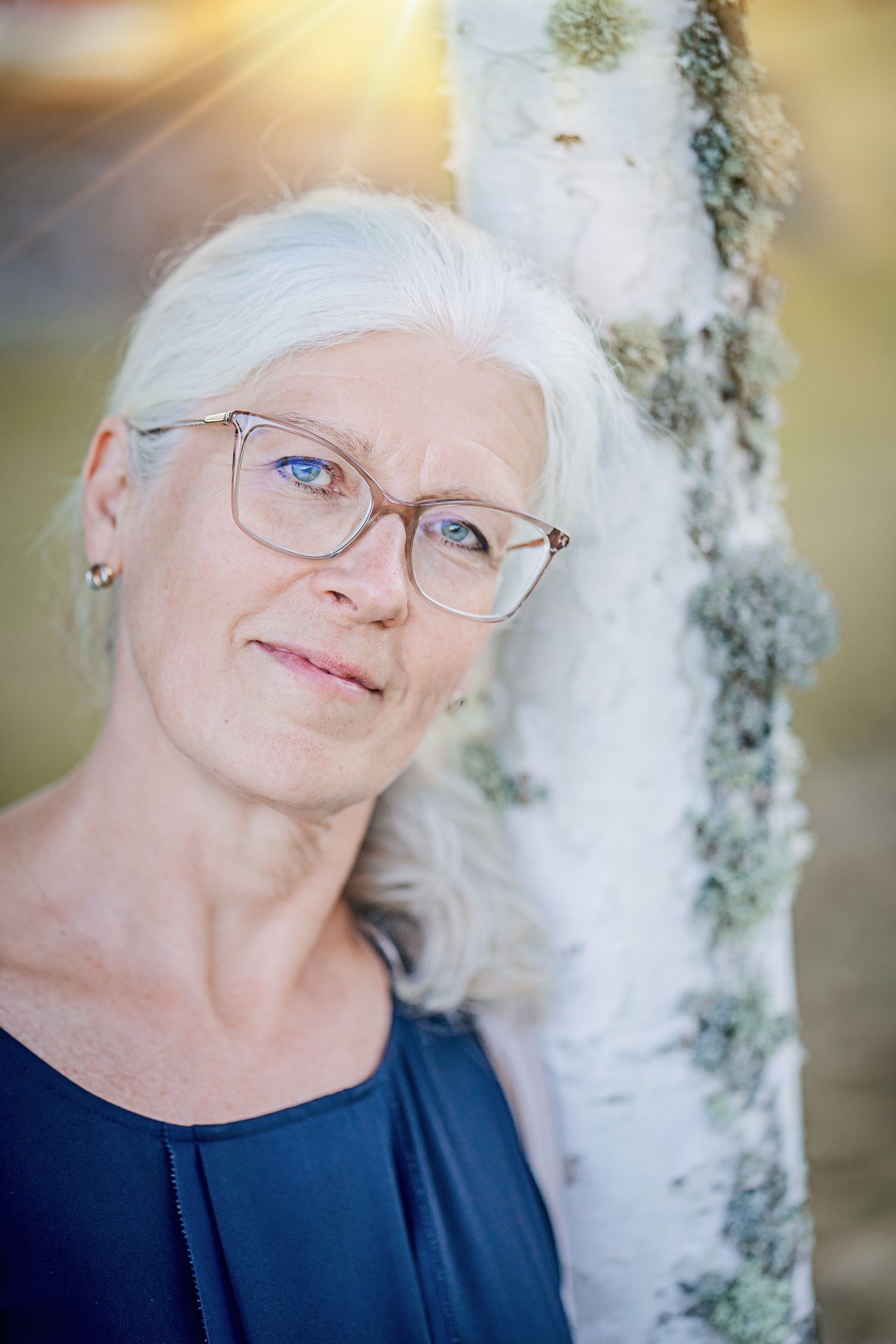 Elegant mature woman with silver hair and glasses posed near a birch tree in natural lighting.