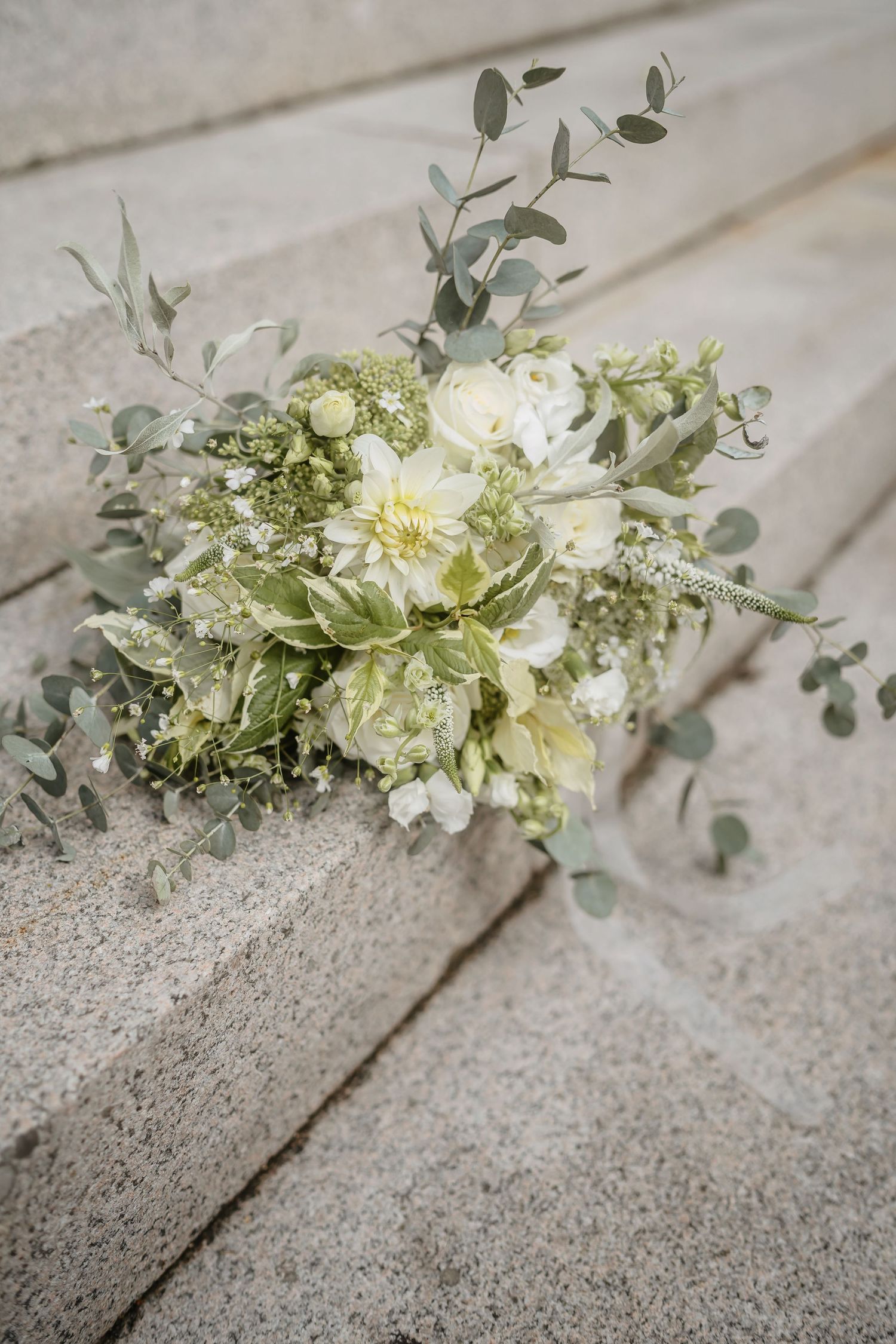 A delicate white and green wedding bouquet with eucalyptus leaves rests on light gray concrete steps.
