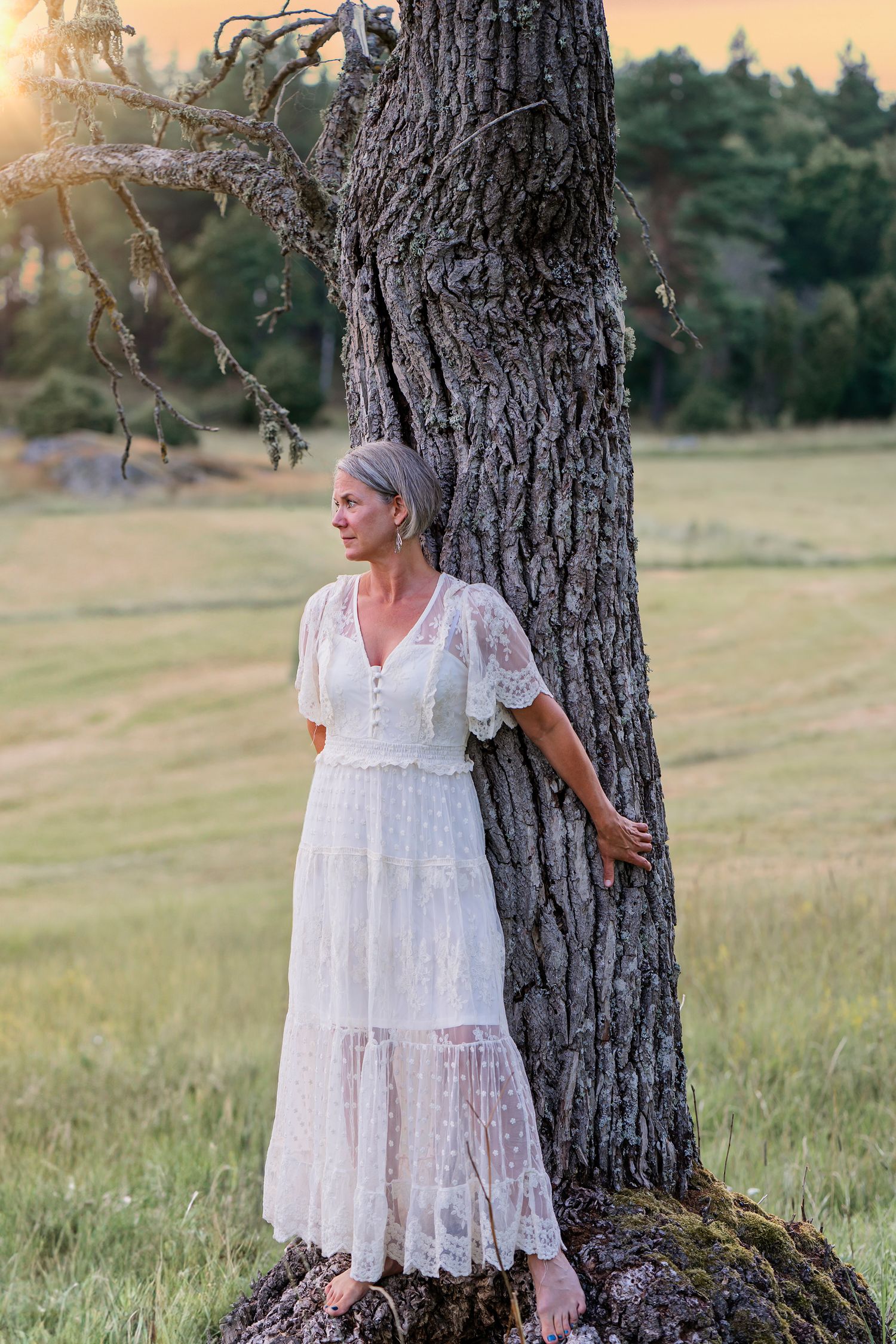 Woman in flowing white lace dress poses gracefully against large tree trunk at sunset.