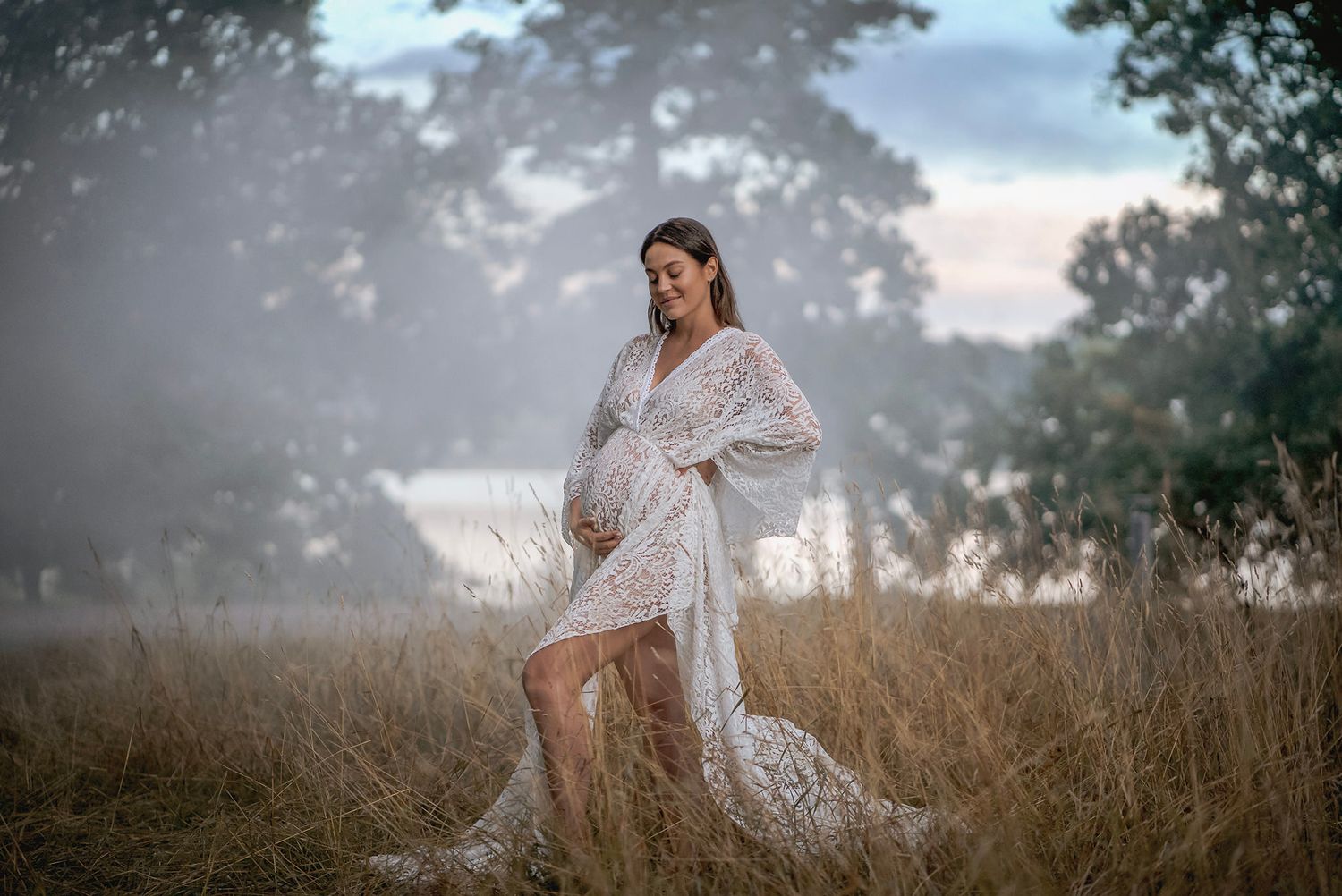 Dreamy maternity portrait of woman in white lace dress standing in misty field at sunset.