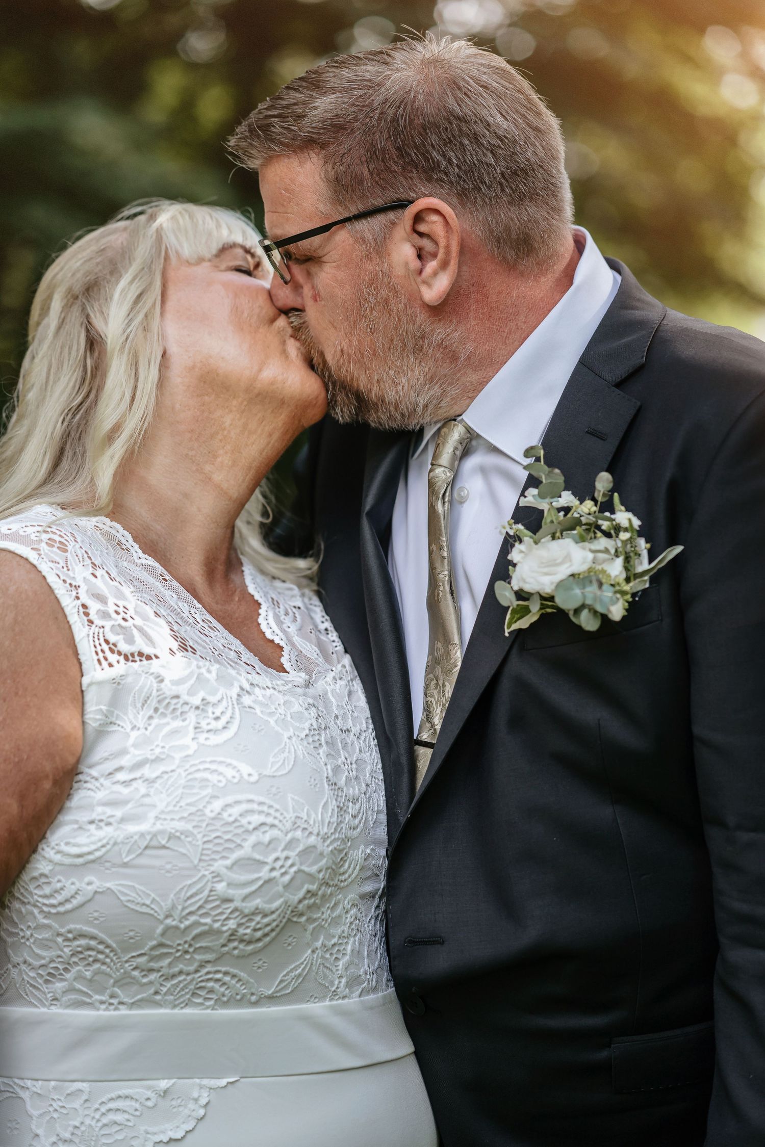A romantic wedding moment captured outdoors with a couple sharing a kiss, wearing white lace and black suit with boutonniere.