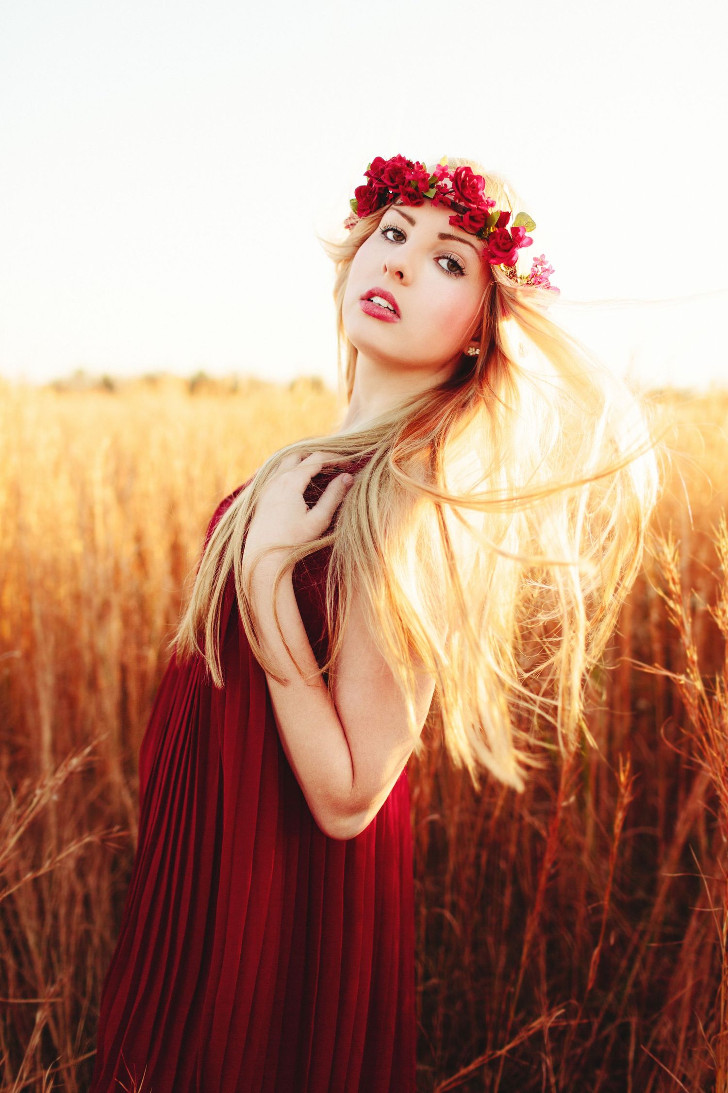 A dreamy portrait in a golden wheat field at sunset features flowing blonde hair, red dress and floral crown against warm light.