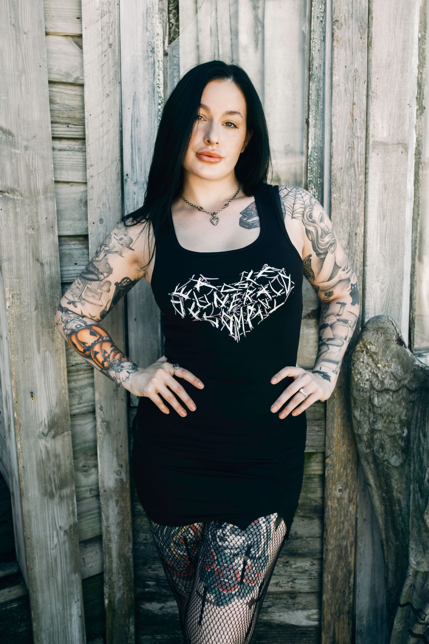 Alternative style model in black dress with arm tattoos poses against rustic wooden wall.