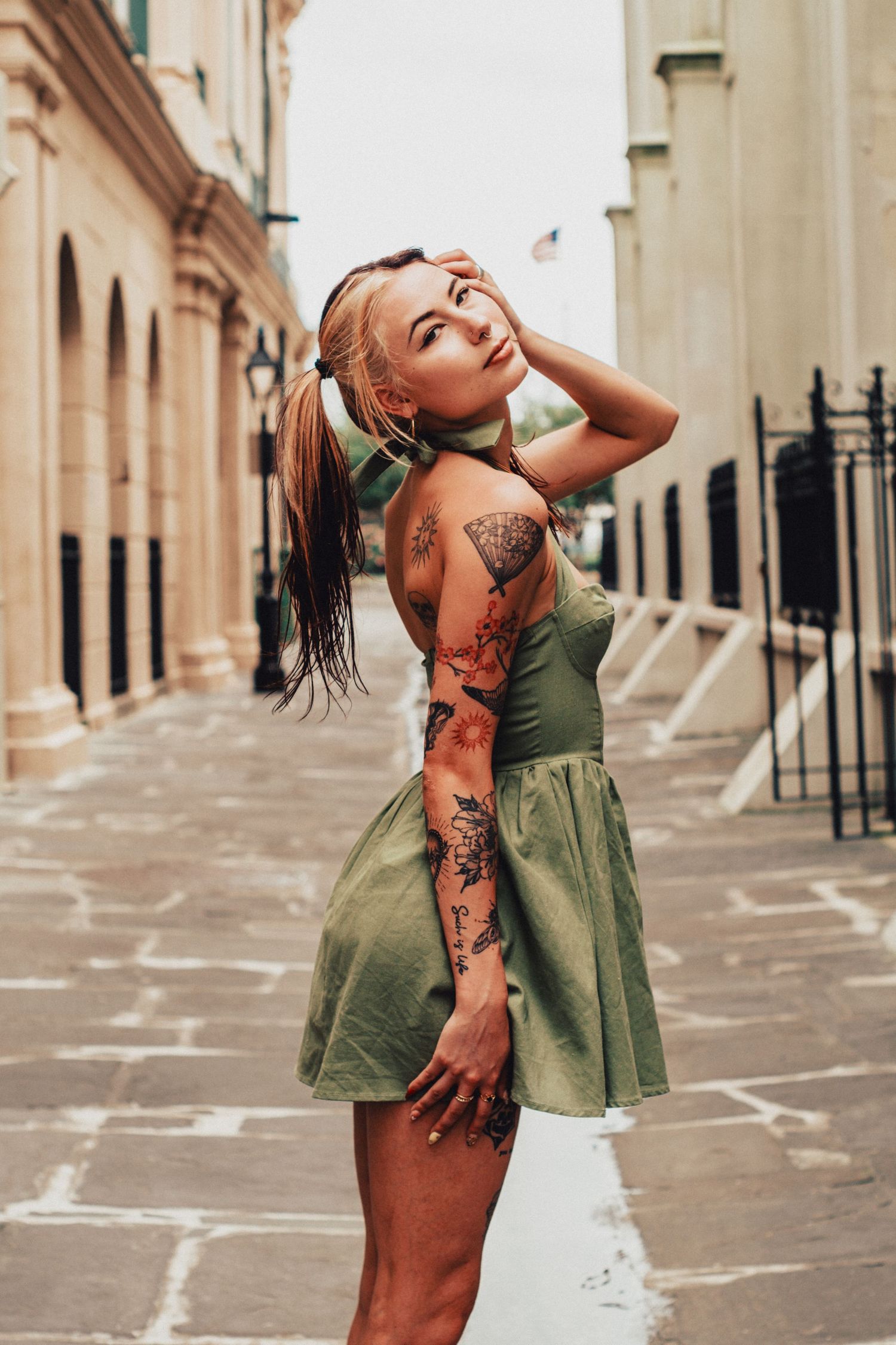 A woman in a green dress with colorful sleeve tattoos poses against a stone wall in an urban alley.