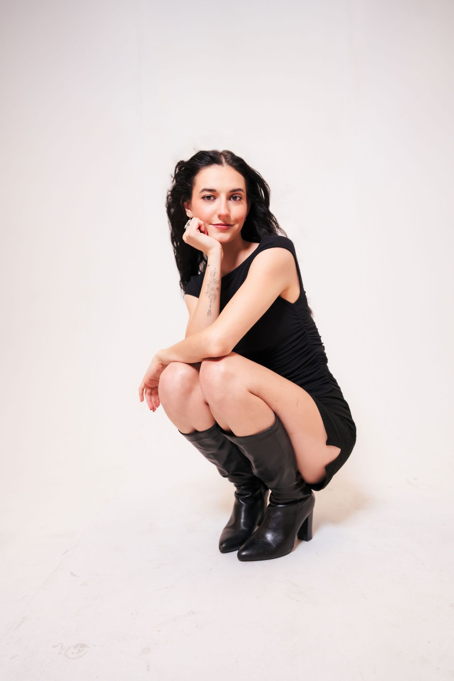 Woman in a little black dress and ankle boots crouches on white background in a stylish pose.