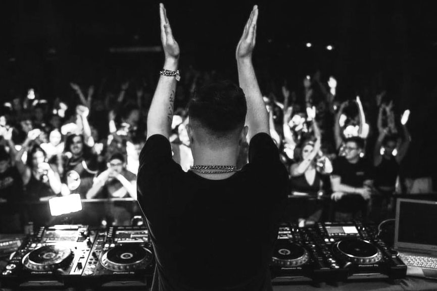 Black and white photo of a DJ performing at a concert with raised hands facing a large energetic crowd.