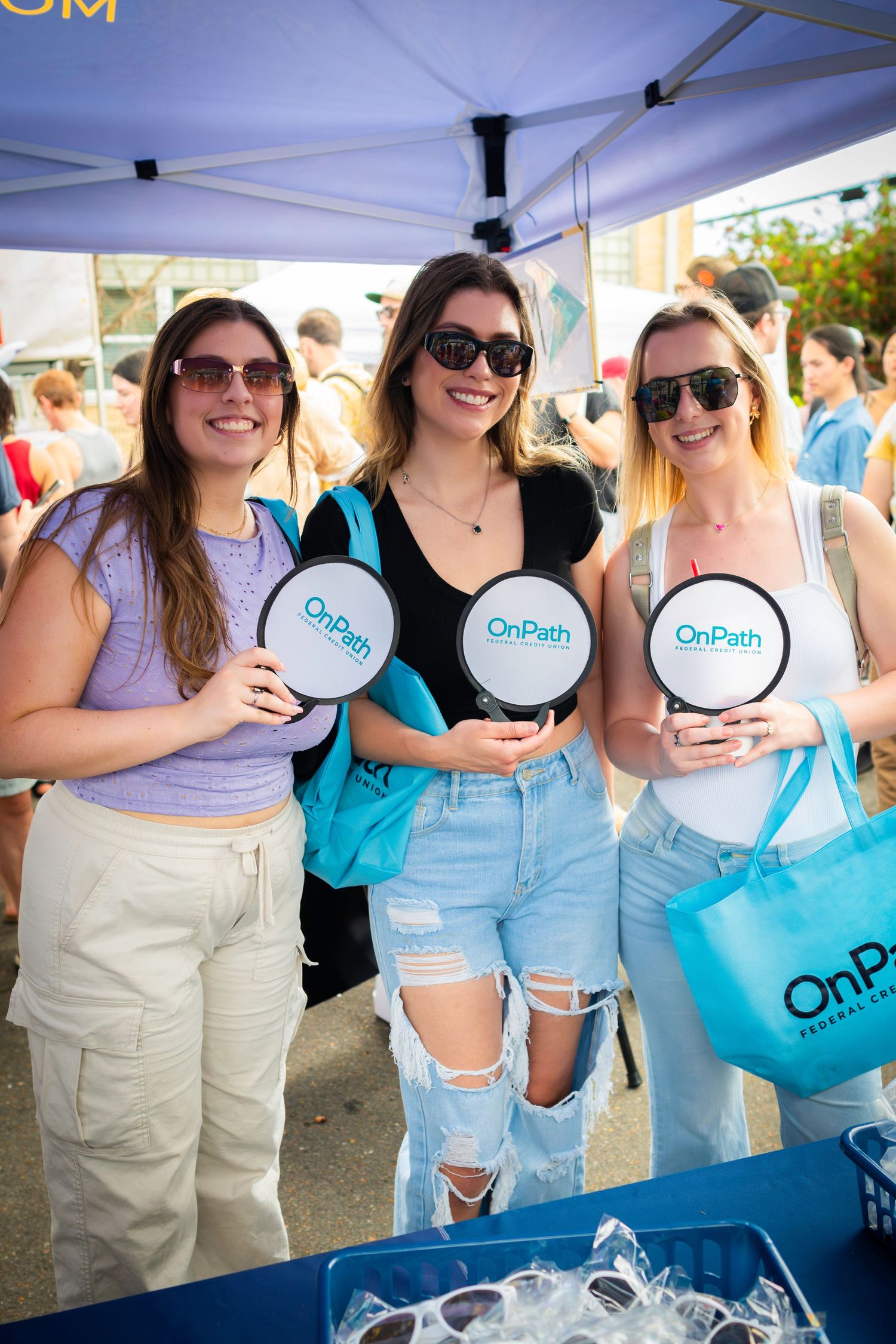 Three friends holding promotional fans at outdoor event under white tent.