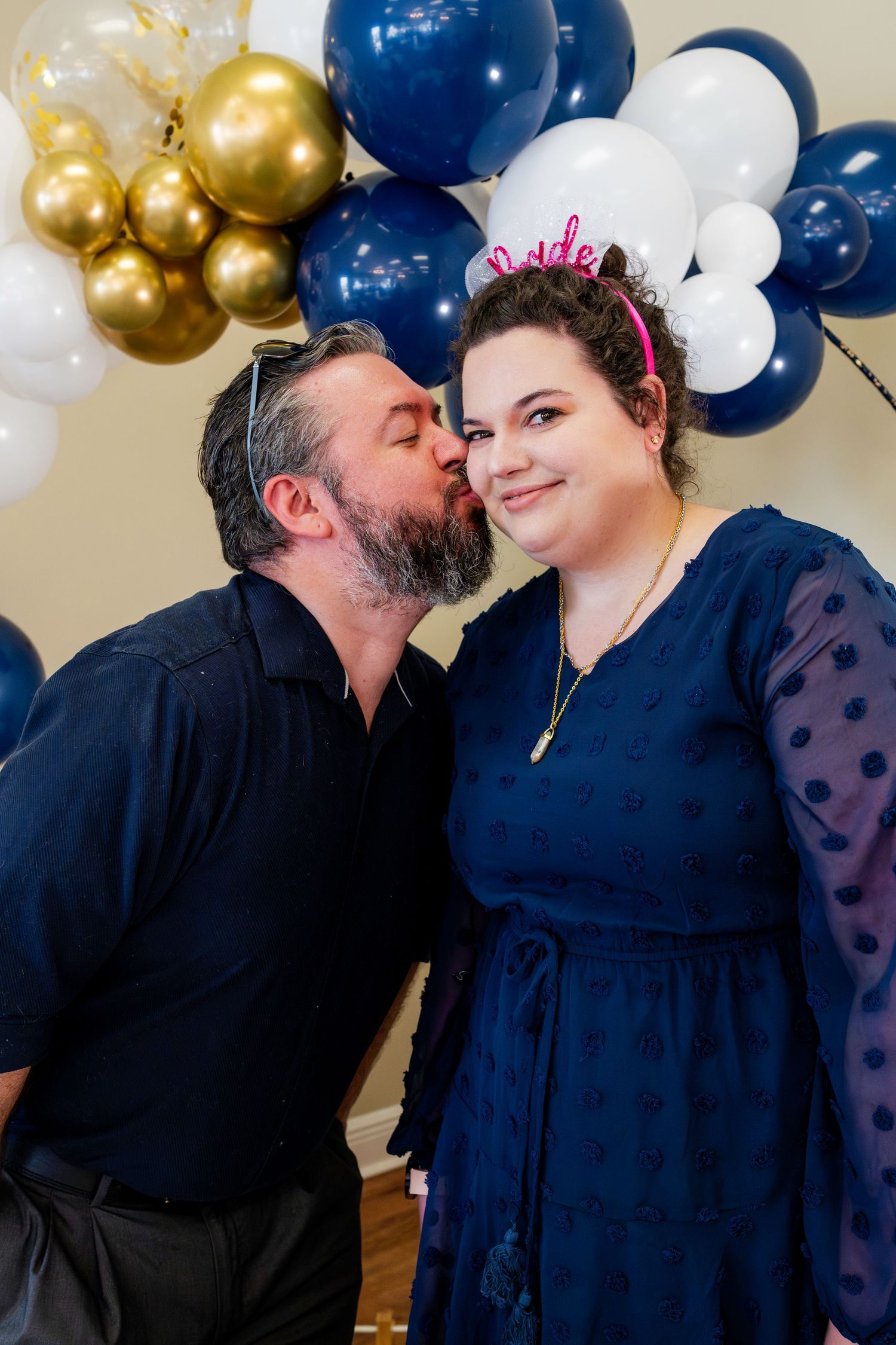 A couple sharing a tender moment in front of blue, white and gold balloon decorations at a celebration.