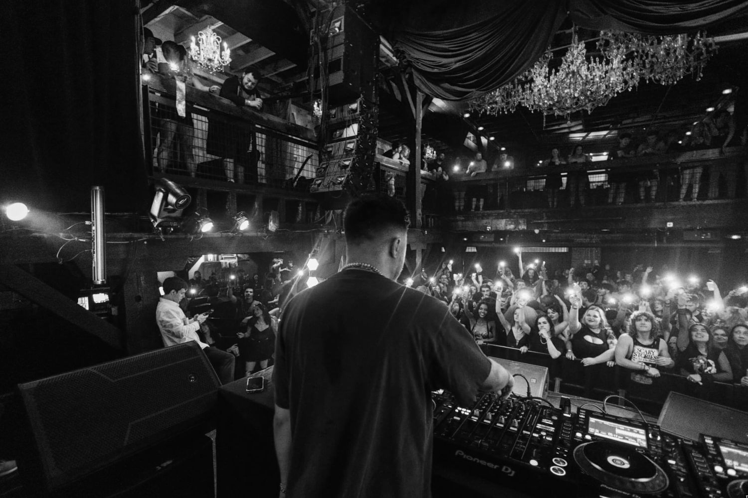 Black and white image of concert venue with chandelier and crowd illuminated by stage lights.