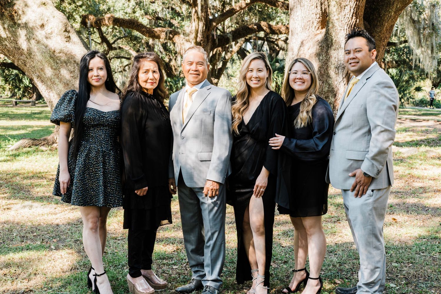 Group photo of elegantly dressed people posing together under oak trees.