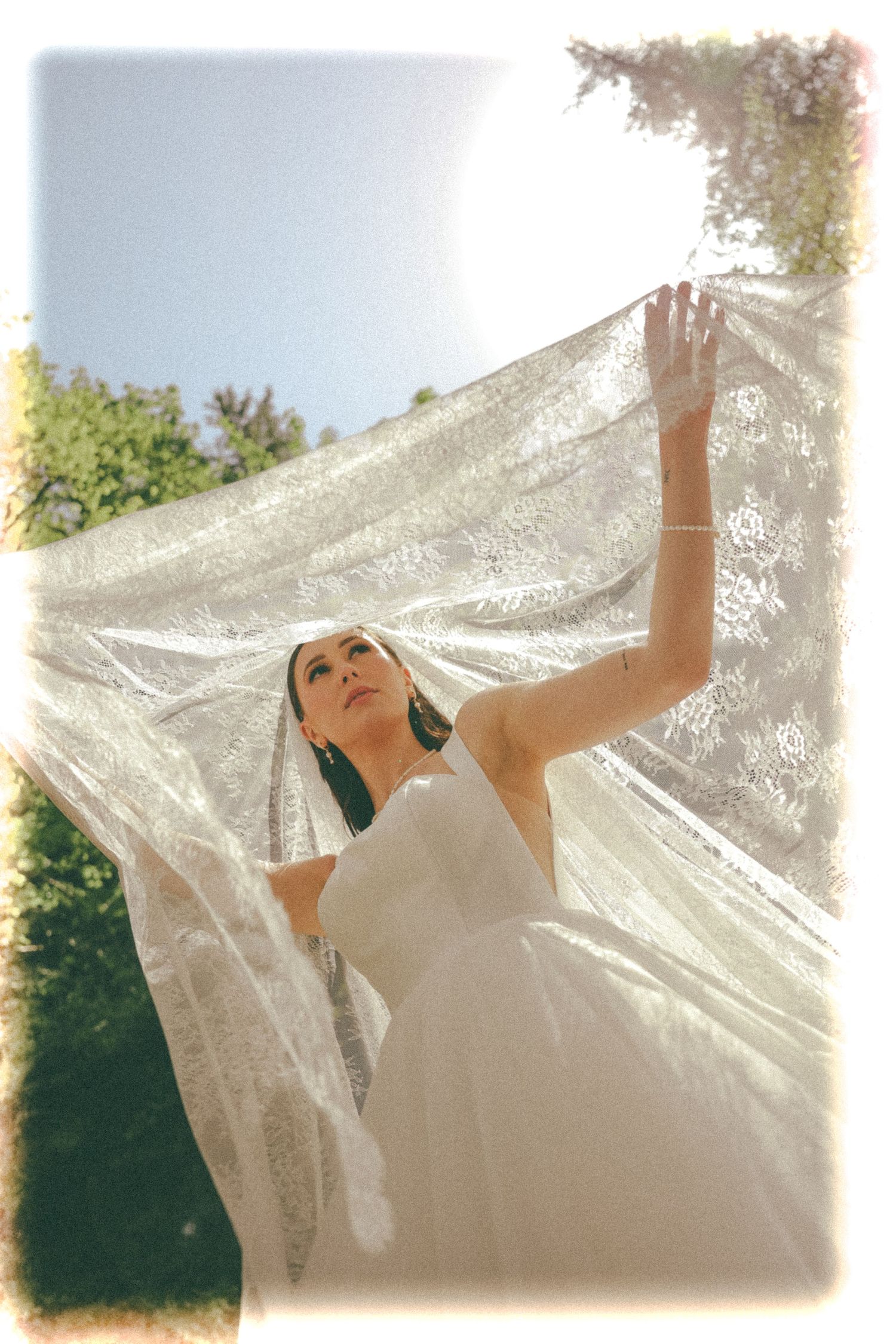 An ethereal bridal portrait with flowing lace veil backlit by sunlight in a garden setting.