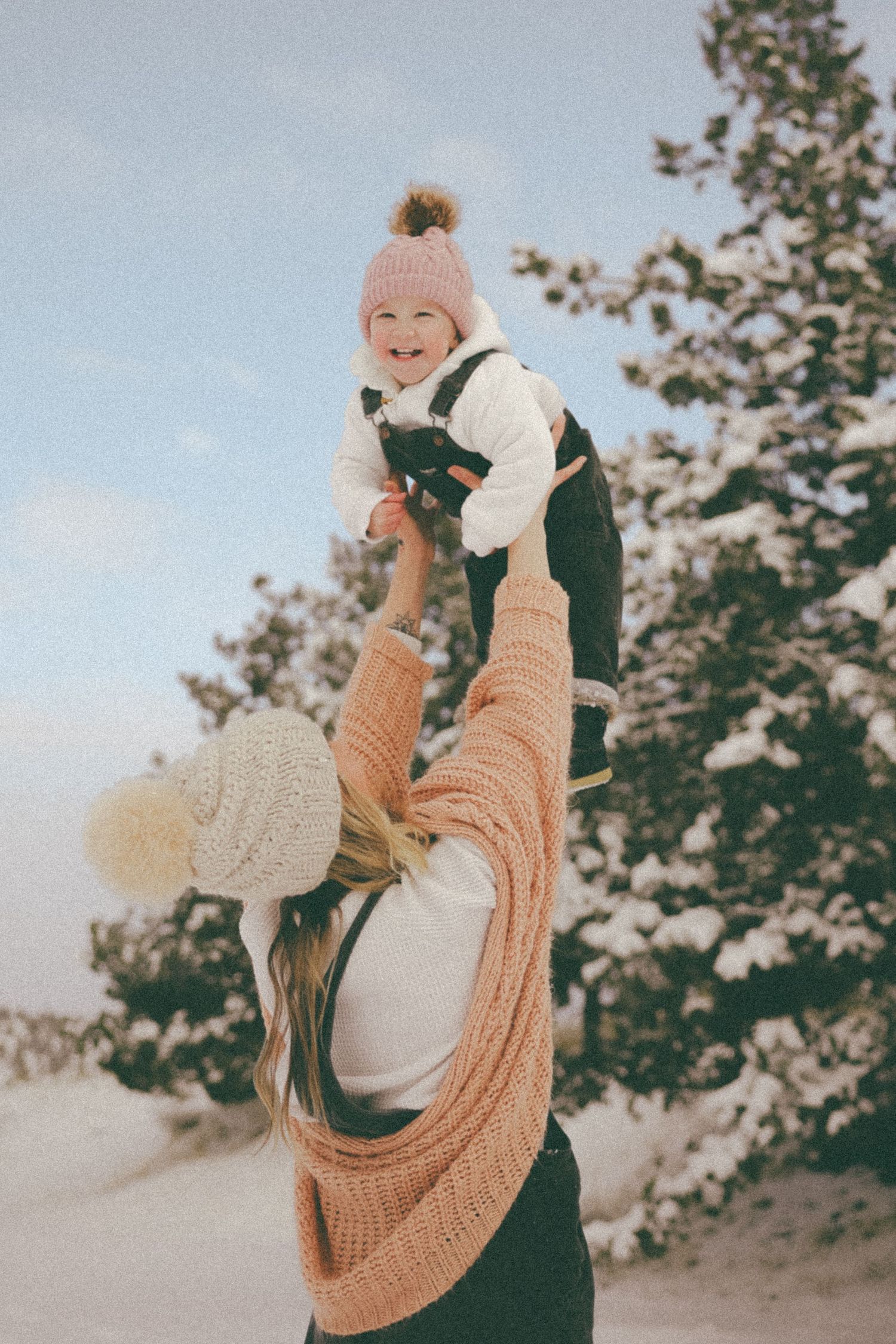 A joyful baby wearing a pom pom hat is lifted high in the air against a winter sky.