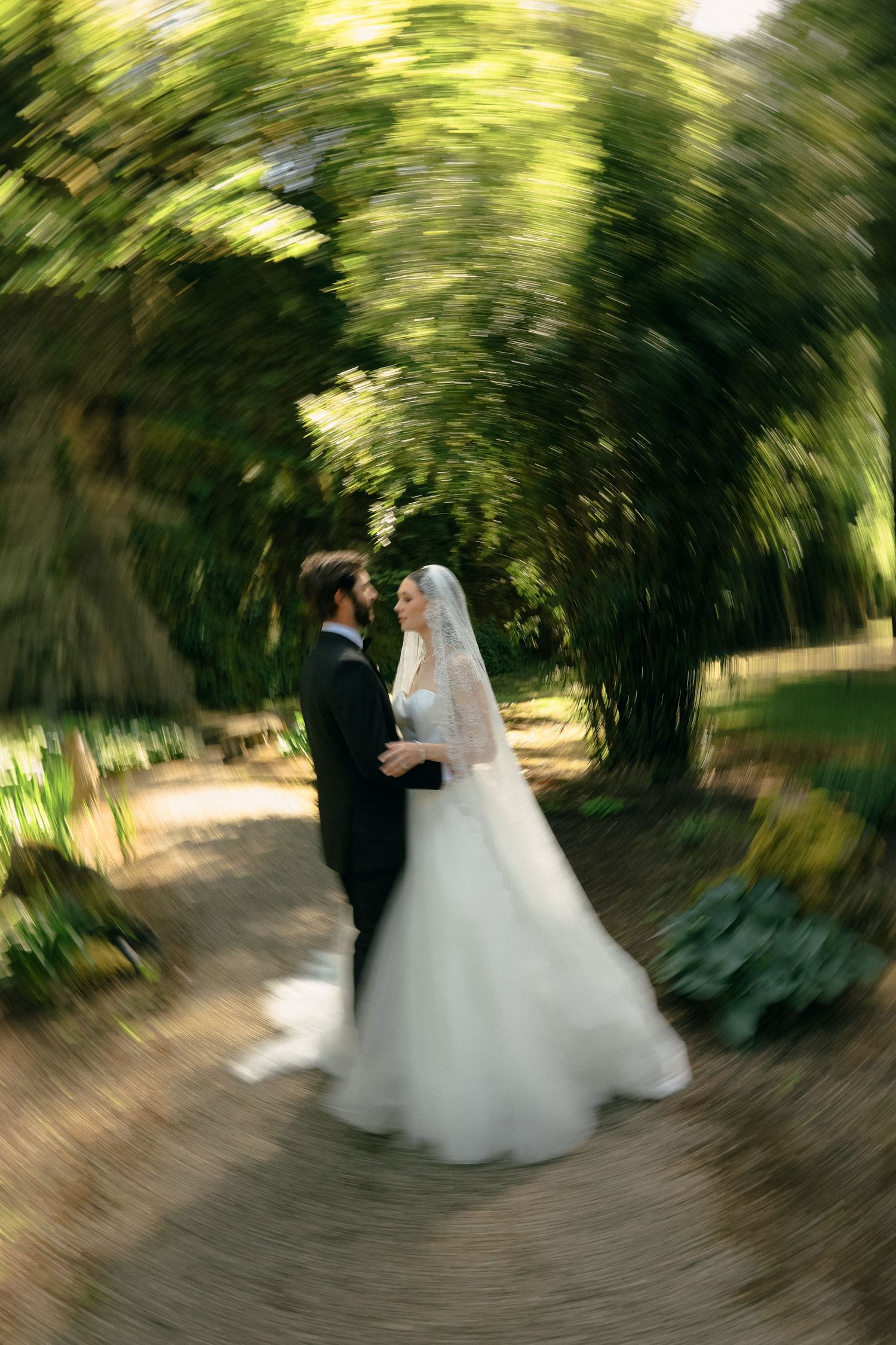 A romantic wedding couple dancing in a swirling motion under a canopy of green trees.