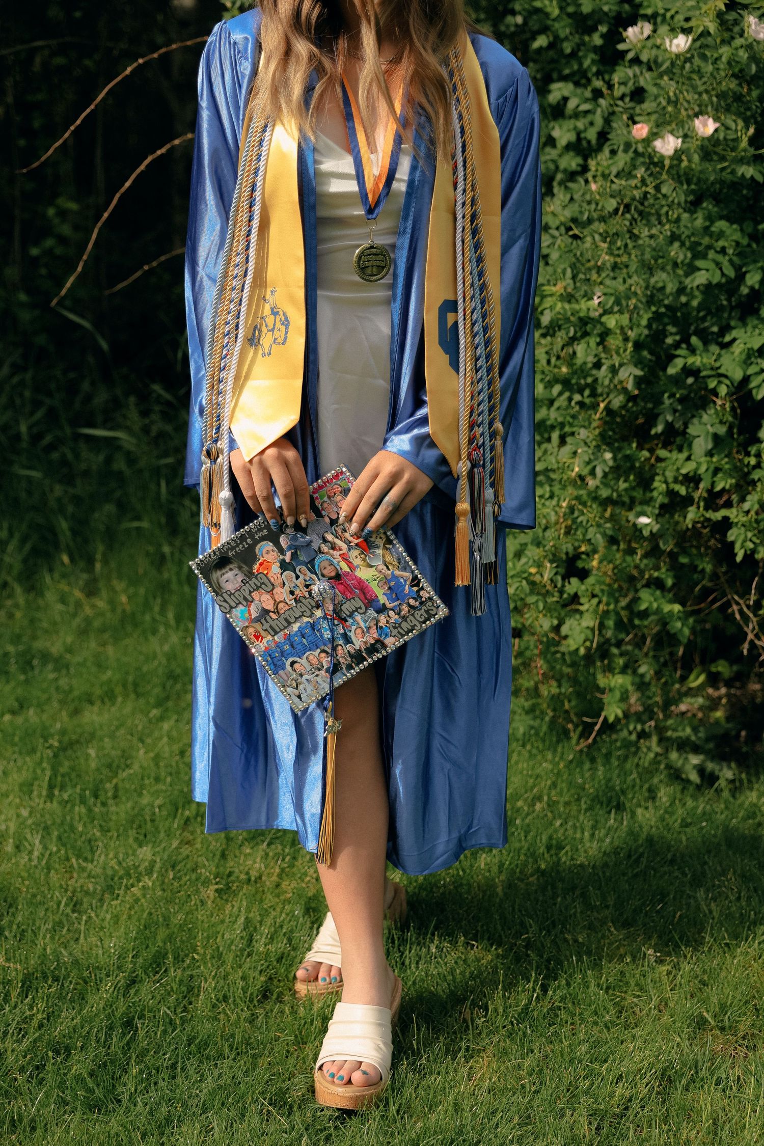 Close-up of graduation gown with decorative handbag and white sandals against green foliage.