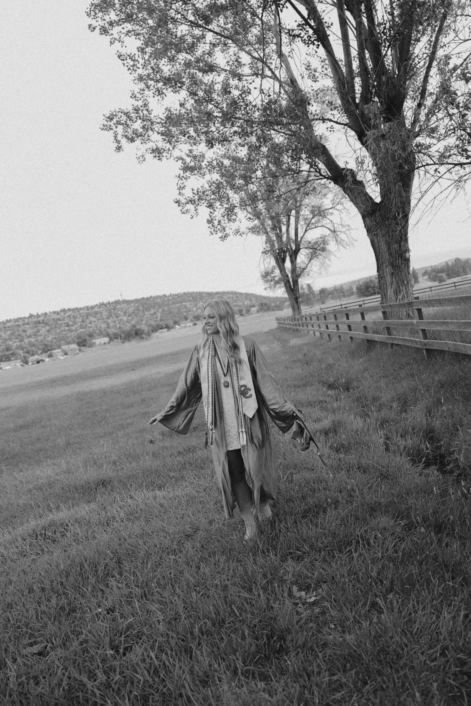 A person stands in a flowing coat along a country fence line with tall trees in black and white.