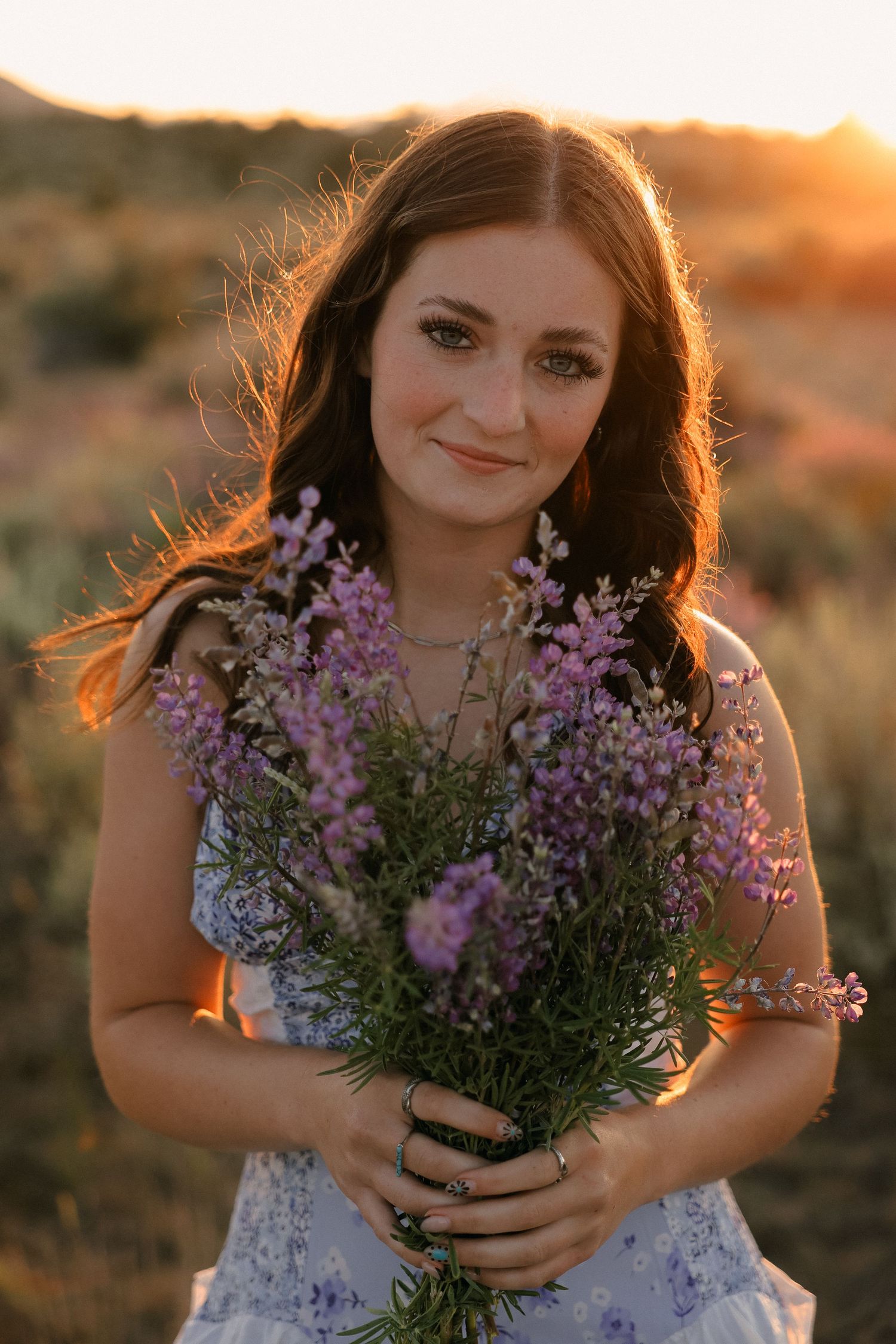 Person holding wildflower bouquet in white dress against sunset backdrop with purple blooms glowing in golden light.