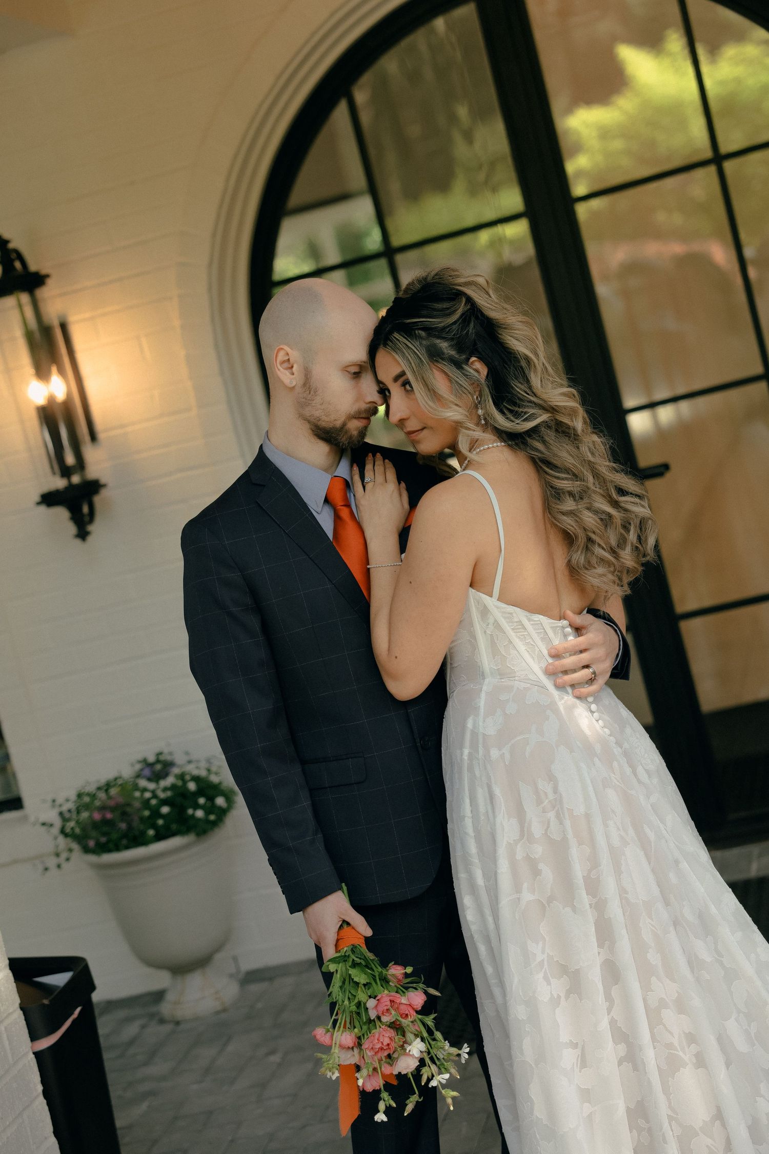 Intimate wedding moment captured between couple in formal attire by elegant arched window.